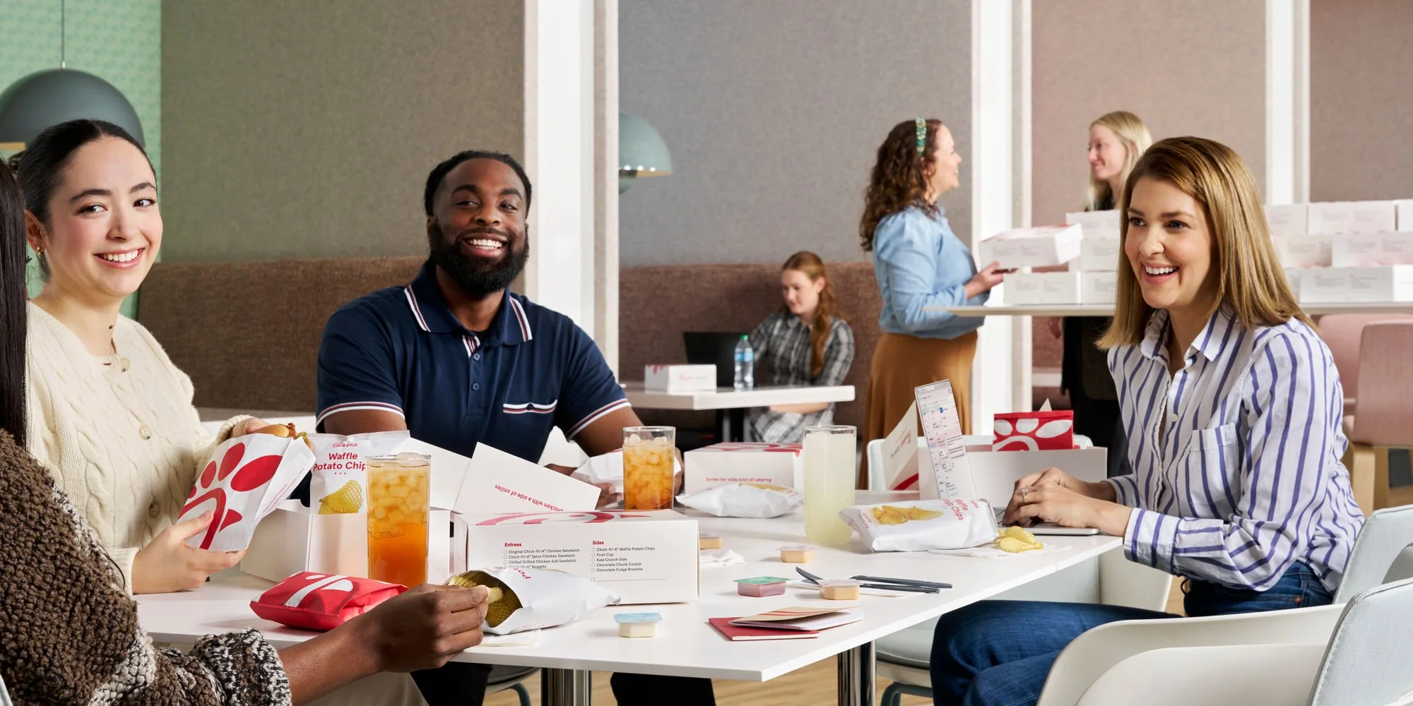 A diverse group of people enjoying a meal at a Chick-fil-A restaurant, smiling and chatting at a white table filled with fast food packaging and drinks.