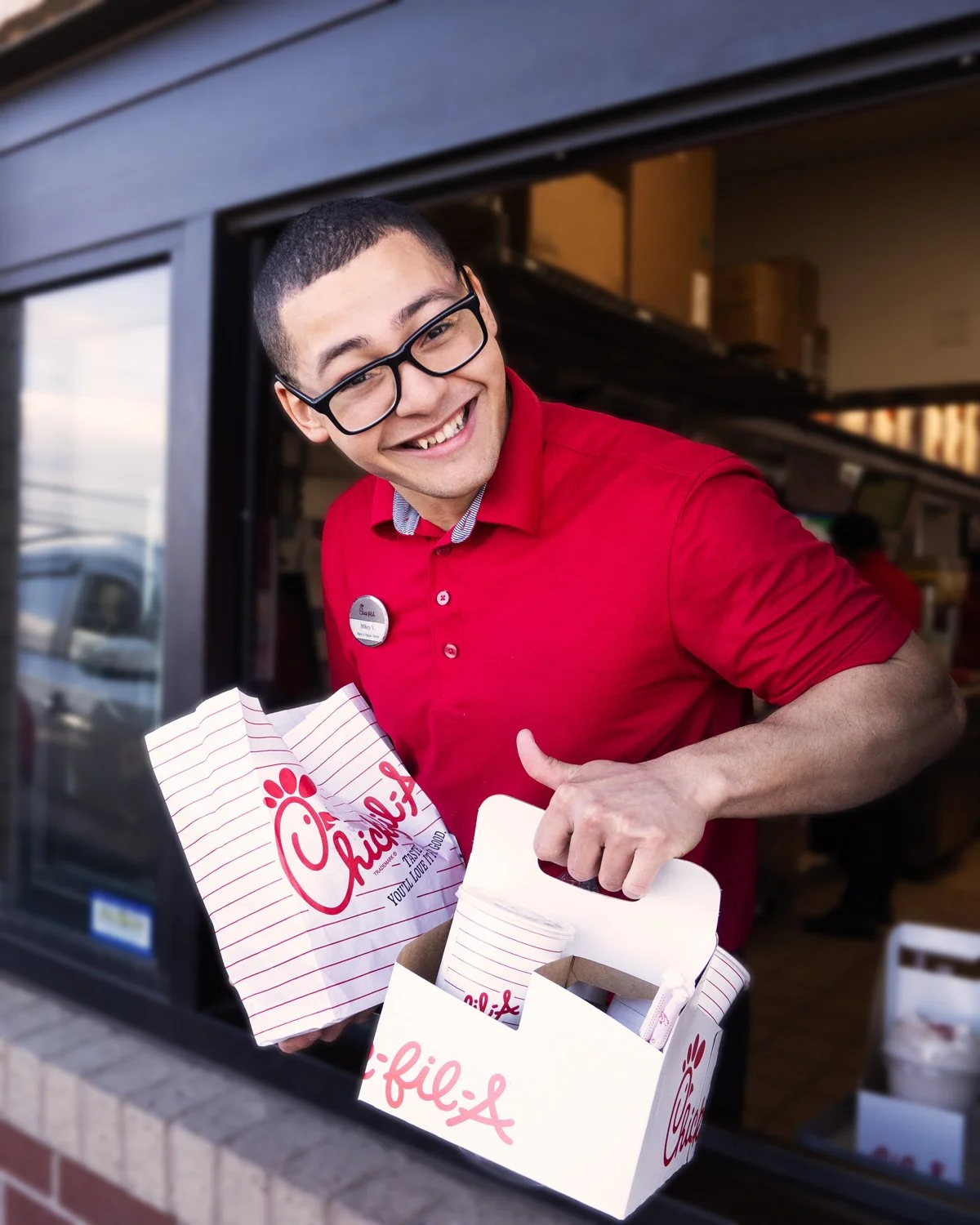 Chick-fil-A employee smiling and handing a boxed meal out of a window, holding a Chick-fil-A bag and a box of food.