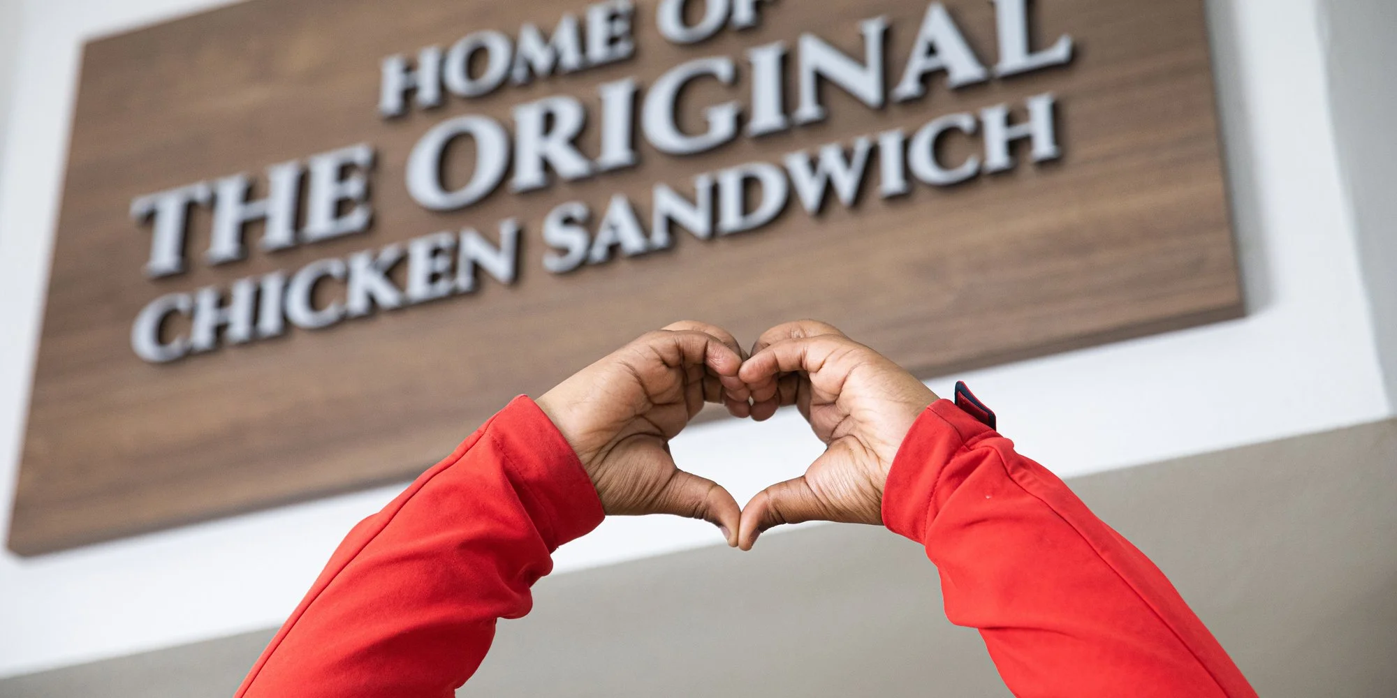 Person wearing a red jacket forming a heart shape with their hands in front of a sign that reads "Home of the Original Chicken Sandwich."