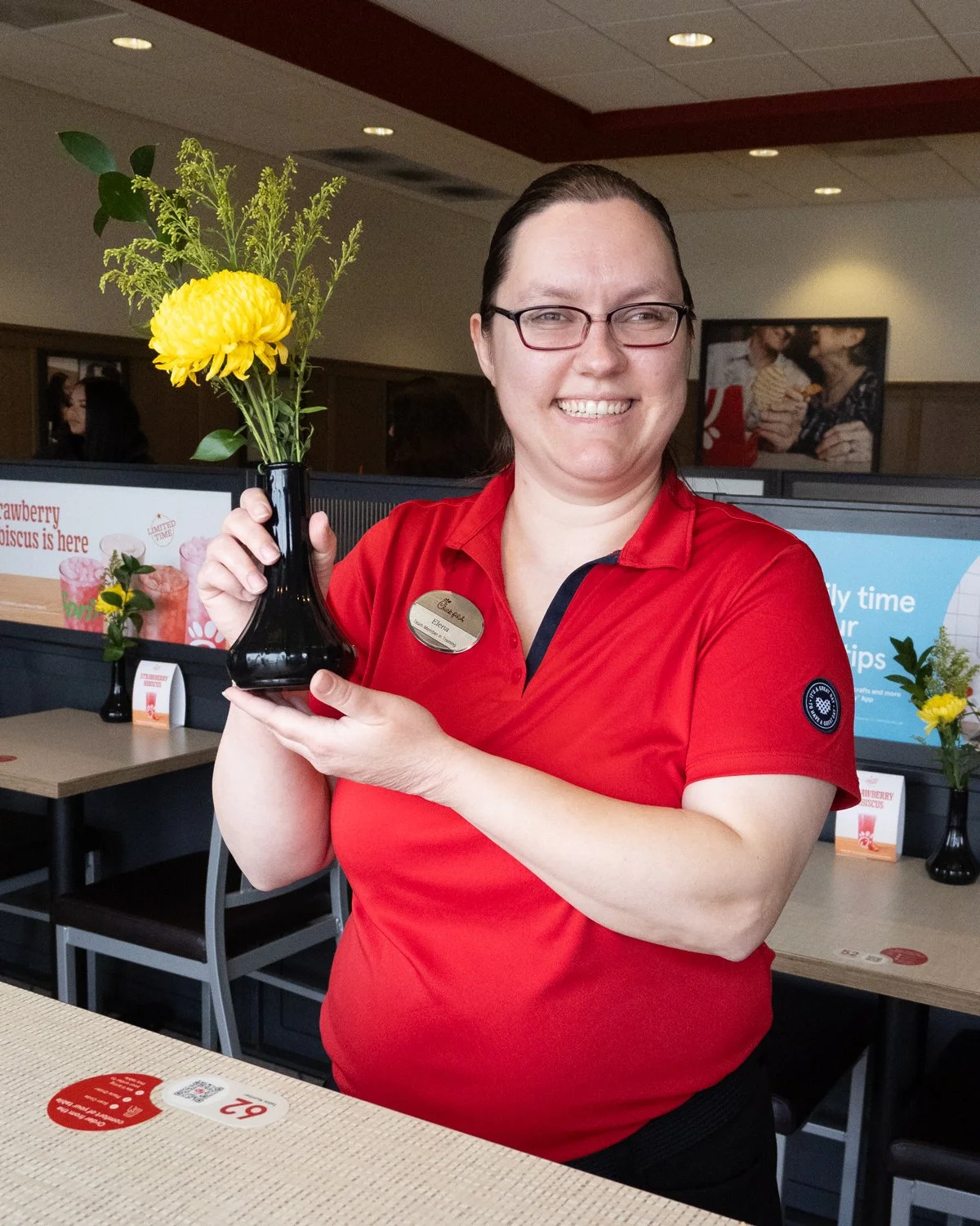 A woman in a red shirt with glasses holds a small black vase with yellow flowers inside. She is smiling inside a restaurant or cafe.