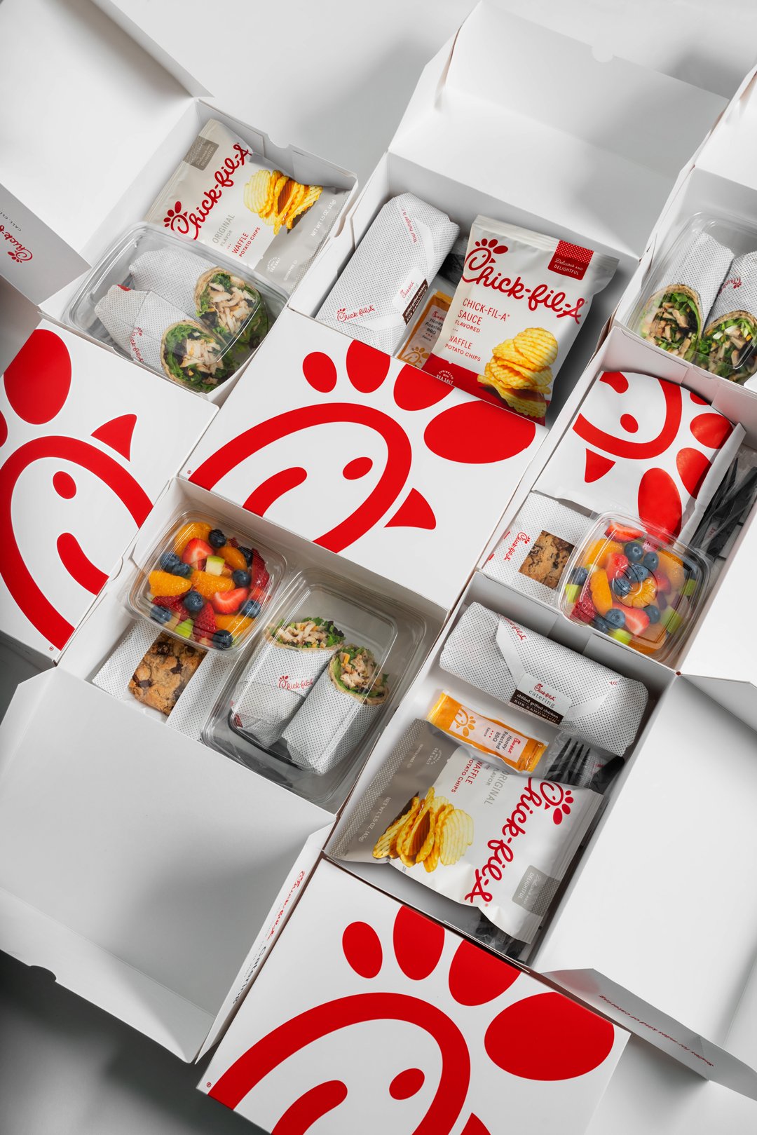 Overhead view of multiple Chick-fil-A lunch boxes and containers filled with sandwiches, fruit, chips, and cookies.