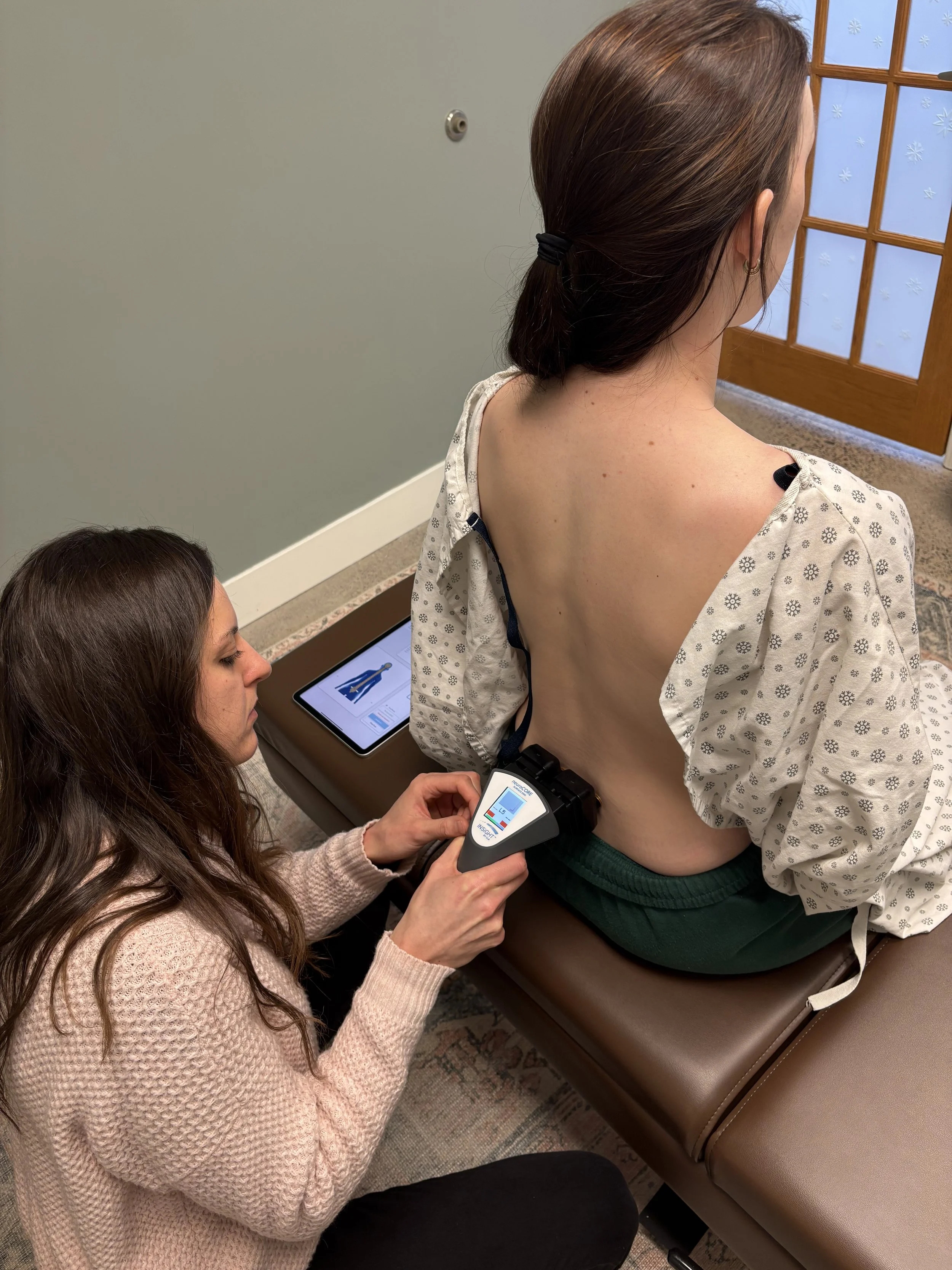 A woman lying on a patient’s back while a healthcare worker uses a medical device on her lower back in a clinical setting with beige walls and a window with snowflake patterns.
