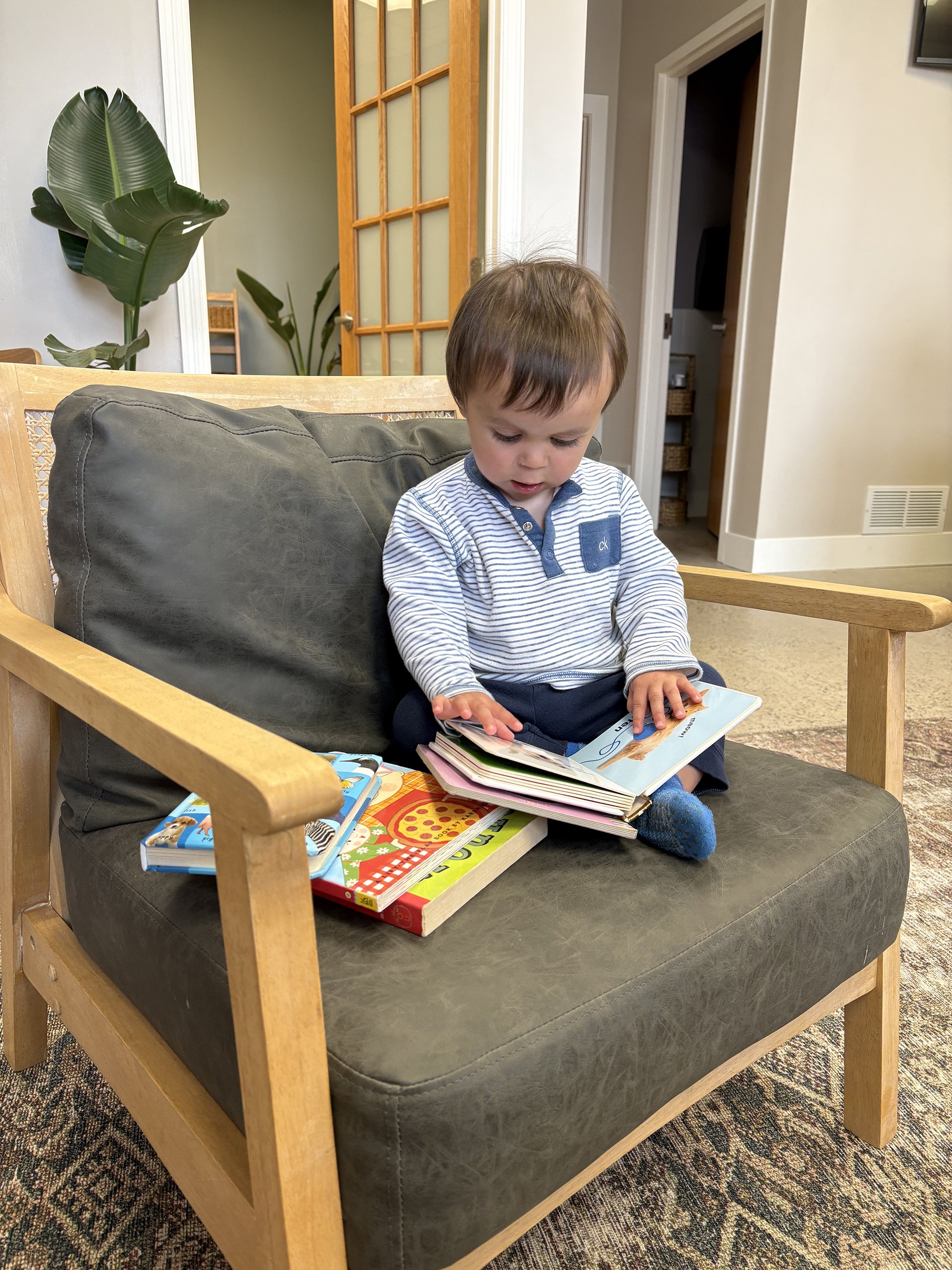 A young boy sitting on a dark green cushioned wooden armchair, looking at a book. Several children's books are placed on the seat beside him. The background shows a room with a doorway, some indoor plants, and a wooden door with glass panels.