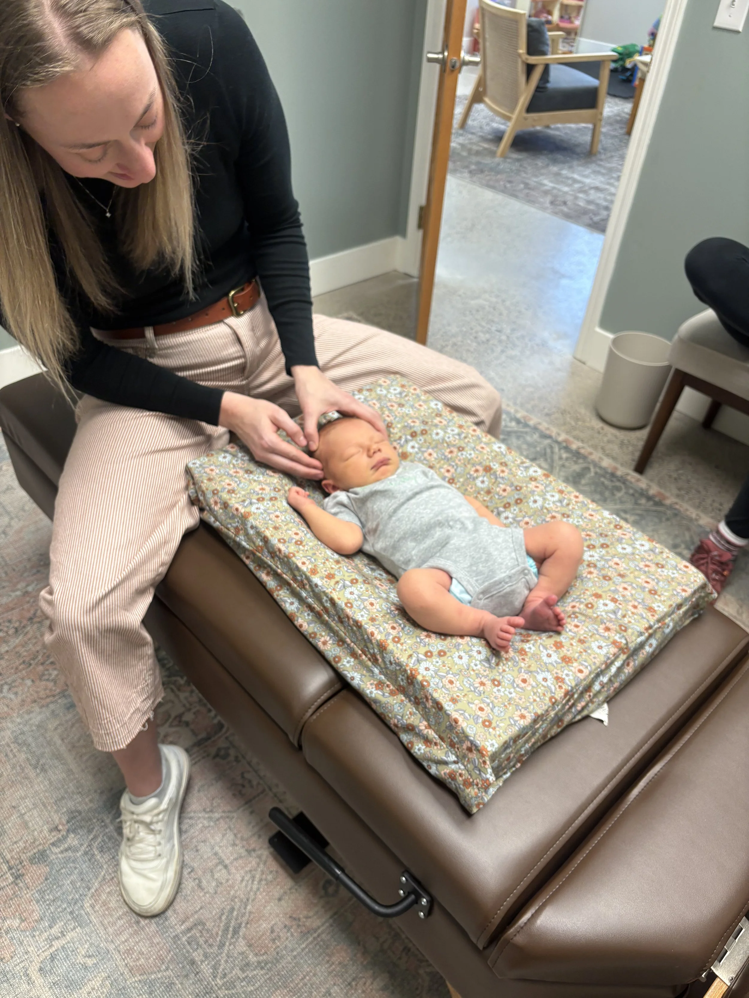 A woman is lying on a padded examination table, receiving an eye check-up from a healthcare provider. The patient is an infant lying on a patterned blanket. The setting appears to be a medical or therapy office with chairs and a hallway in view.