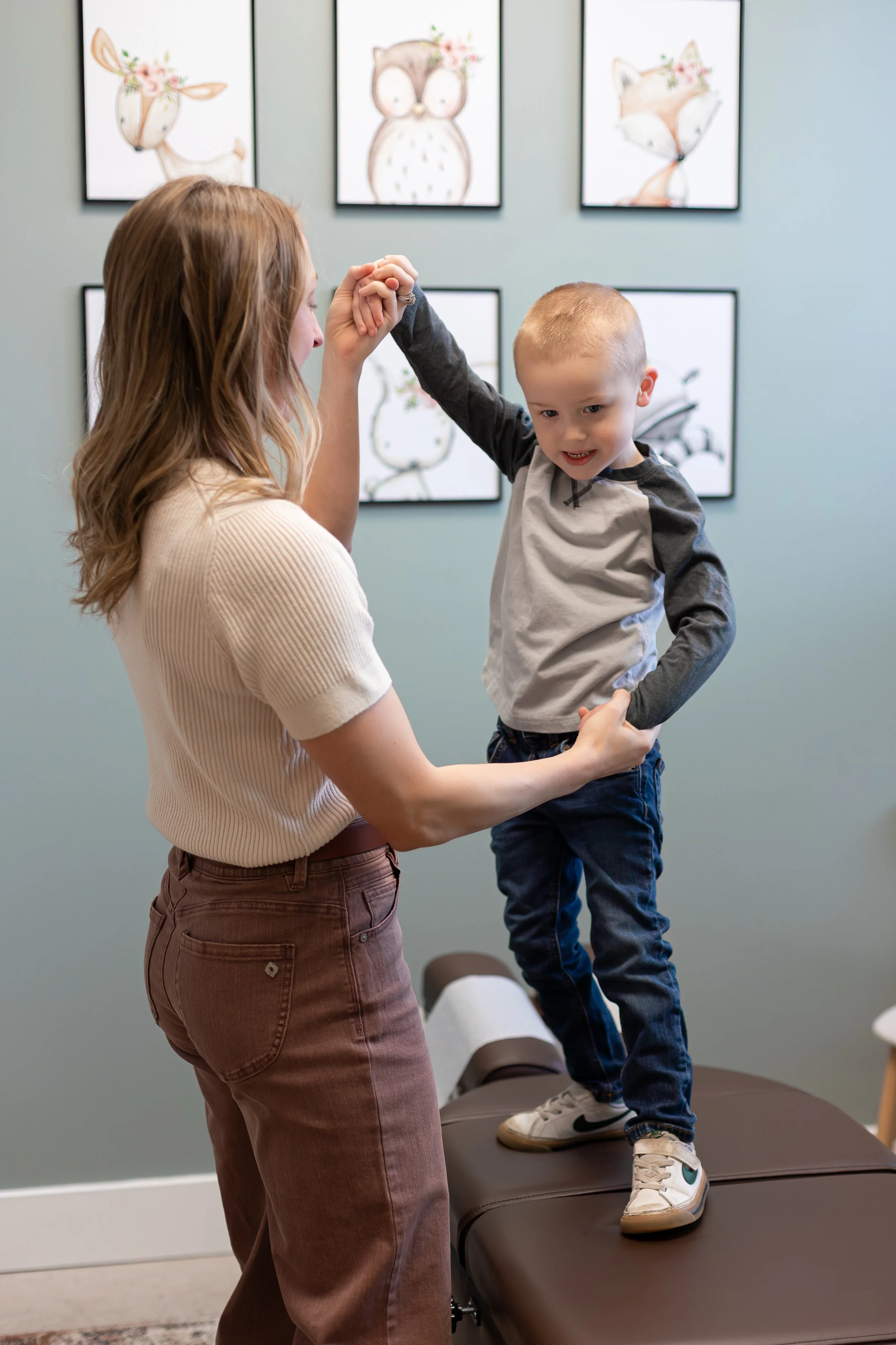 A woman with long, wavy red hair is holding hands with a young boy who is standing on an examination table in a doctor's office or clinic. The boy, dressed in a gray and black long sleeve shirt and dark jeans, is smiling and appears to be engaged in a playful activity. Behind them, a light blue wall is decorated with framed watercolor or pastel illustrations of whimsical animals, including a deer, an owl, and a fox, each with floral accents.