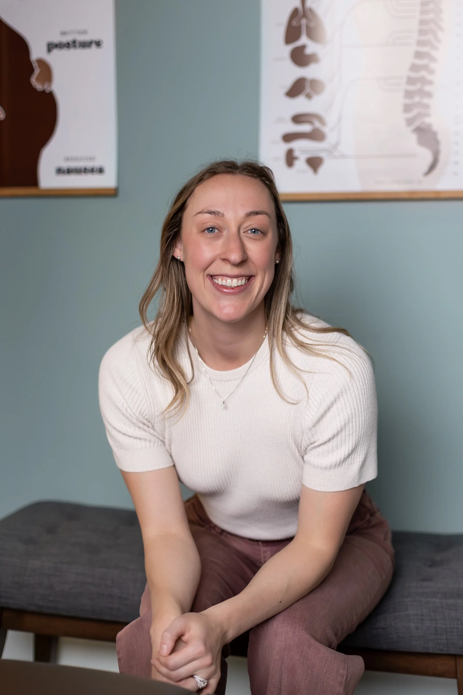 A young woman with long light brown hair, blue eyes, and fair skin, smiling and sitting cross-legged on a bench in front of a light green wall. She wears a cream-colored short-sleeve sweater, maroon pants, a necklace, earrings, and a ring, with posters of anatomical diagrams on the wall behind her.