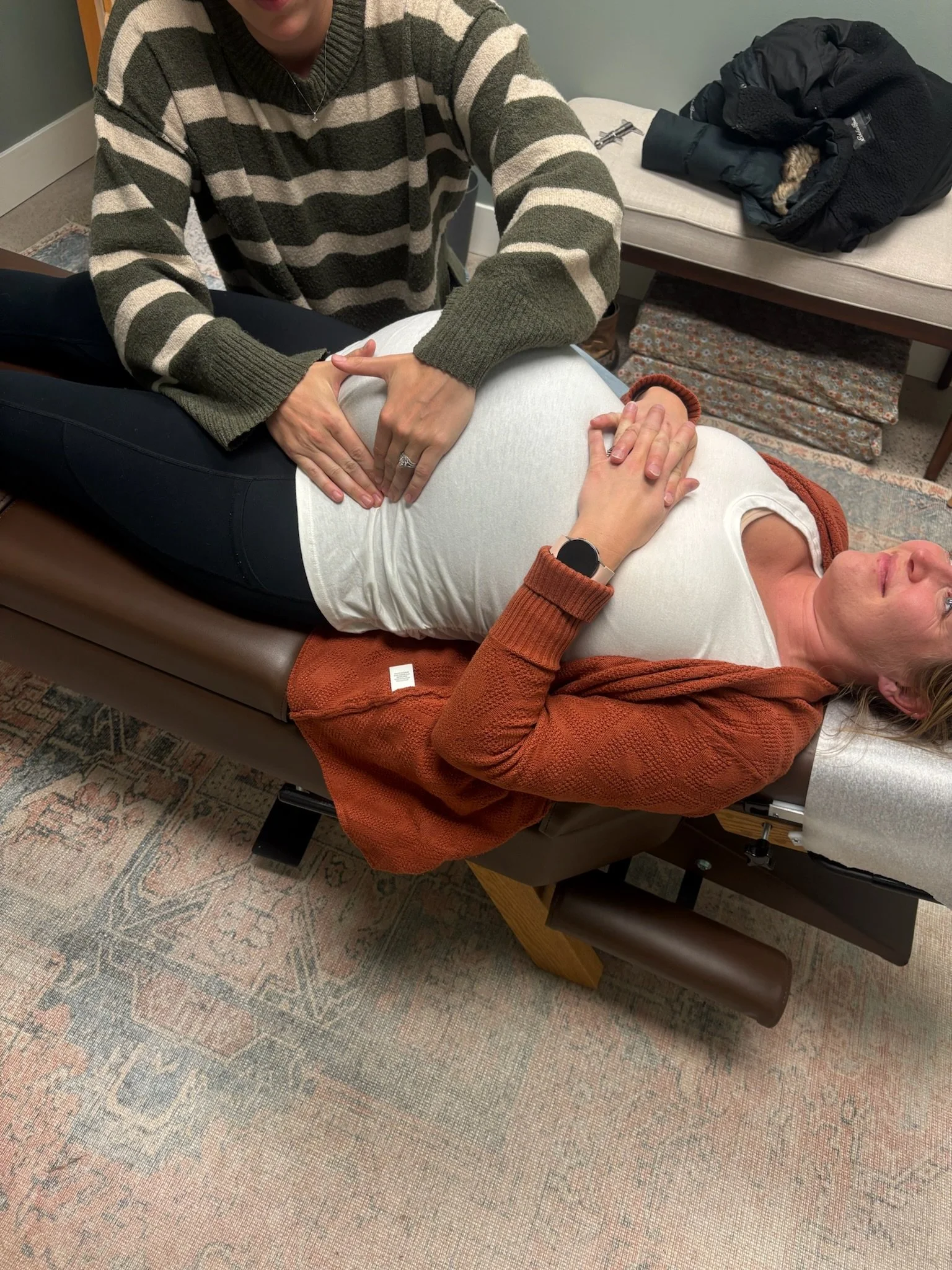 A person lying on a medical examination table during a medical consultation with a healthcare provider who is palpating their abdomen. The healthcare provider is wearing a striped sweater, and the patient is wearing a white shirt and a watch.
