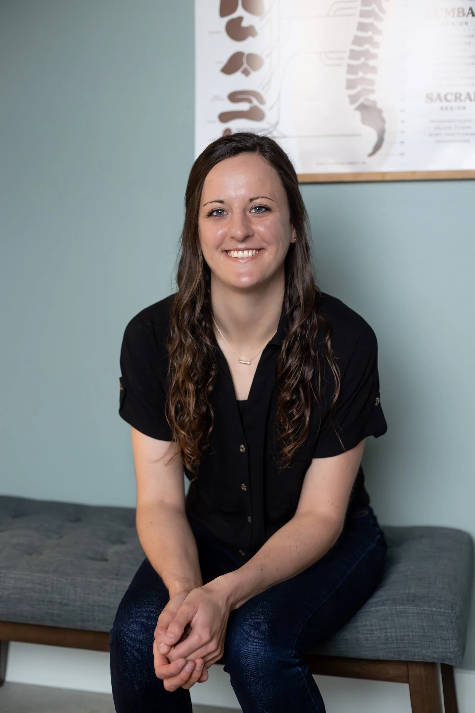 A woman with long wavy hair sitting on a bench in front of a dental poster on the wall. She is smiling and wearing a black shirt and blue jeans.