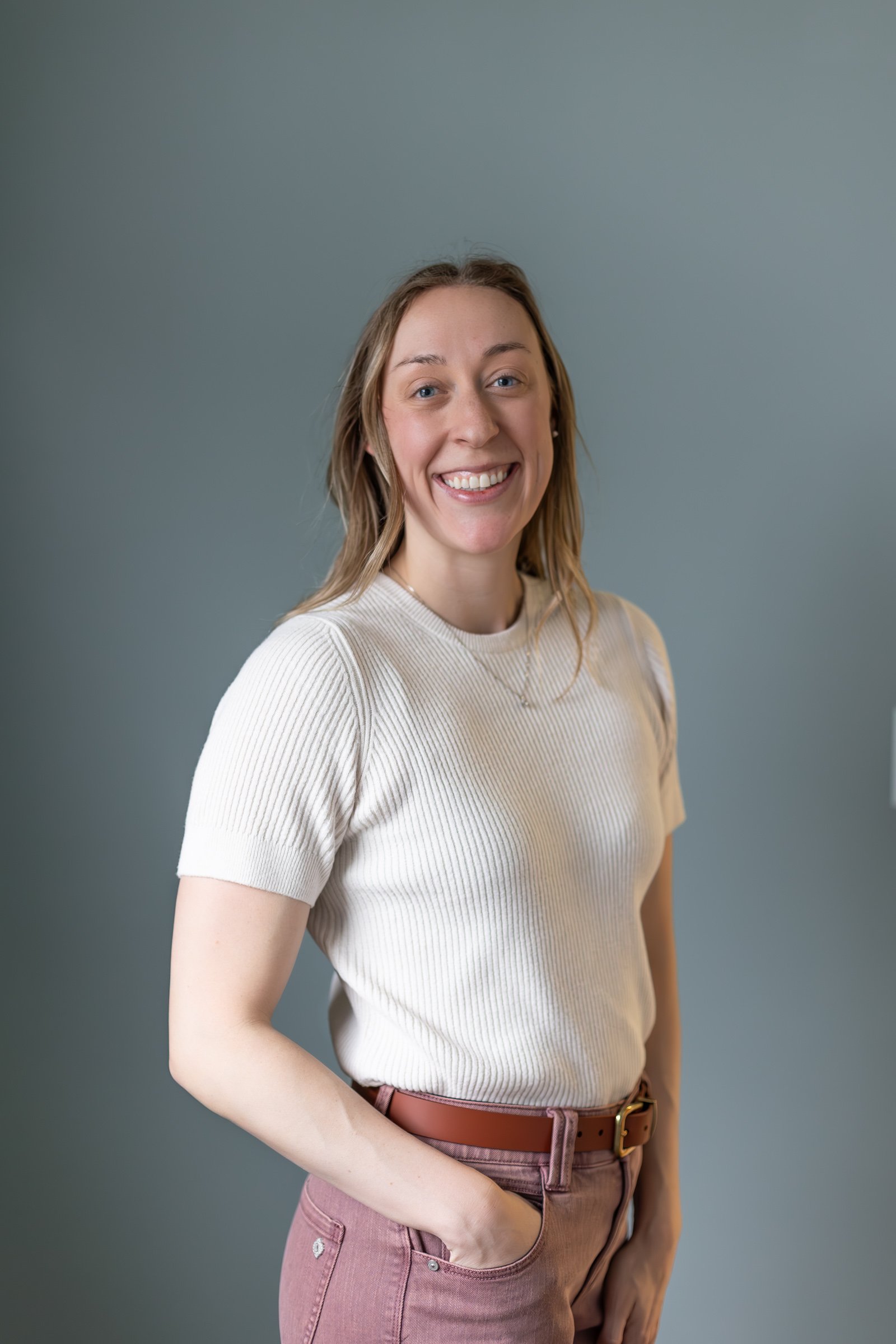 A smiling woman standing against a plain gray background, wearing a light beige ribbed short-sleeve top and pinkish-brown pants with a matching belt.