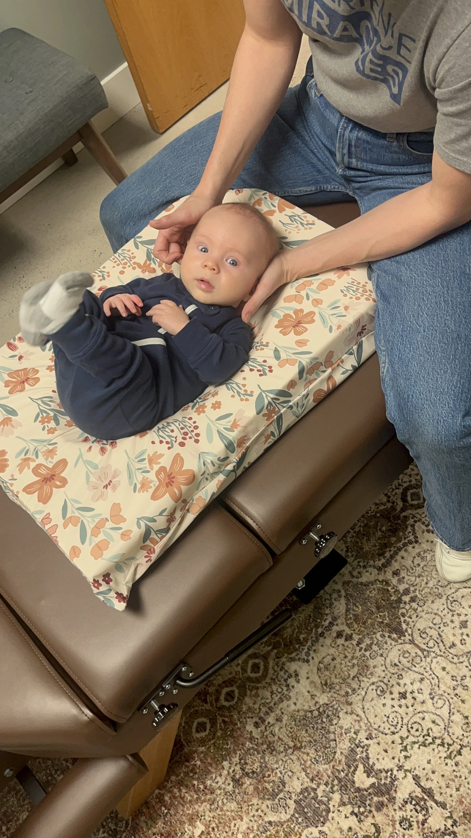 A baby lying on a changing pad with floral pattern, with an adult gently holding their head and legs, in a room with a brown bench, a wooden cabinet, and a patterned rug.