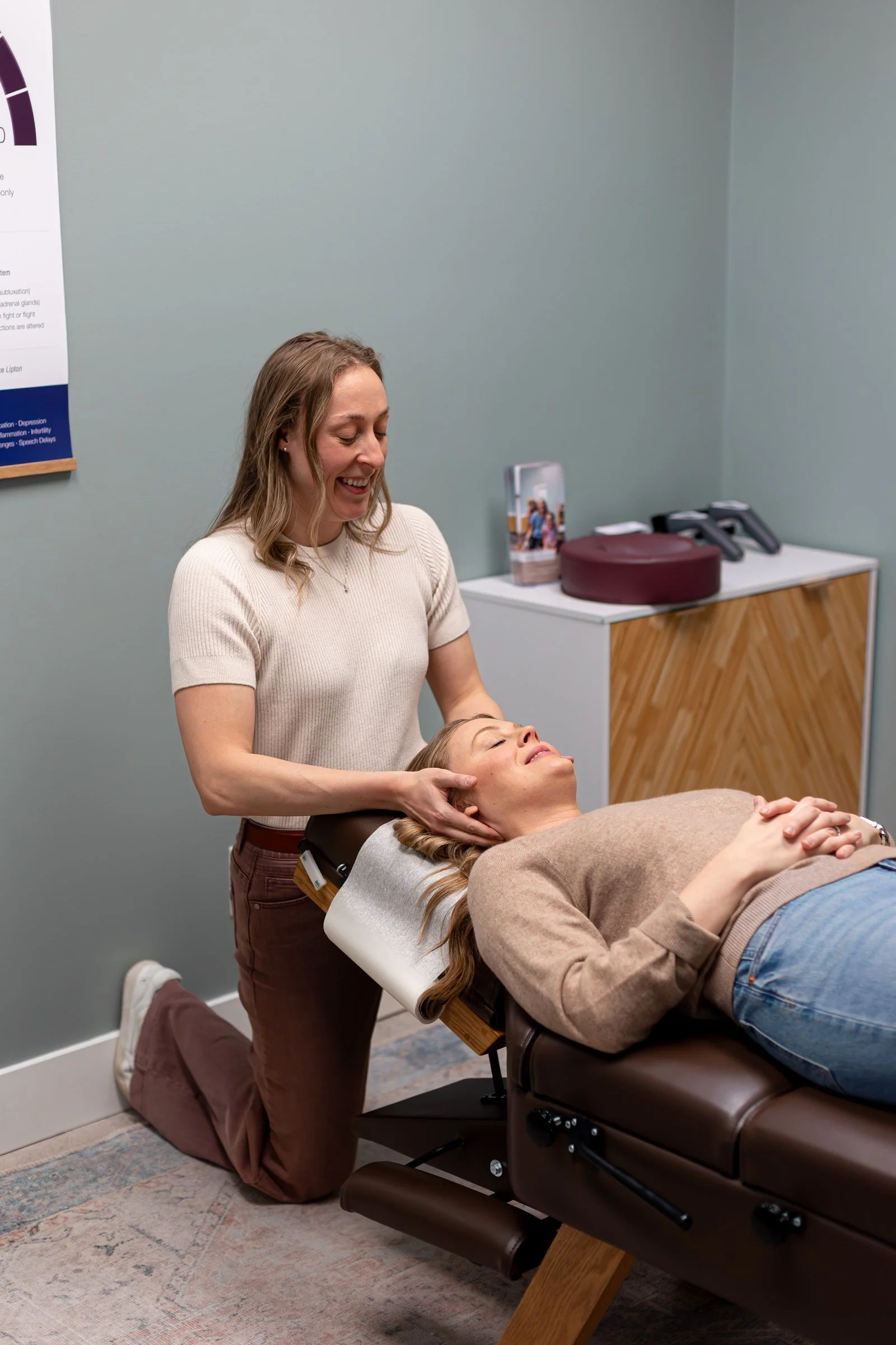 A woman receiving a massage from a therapist in a treatment room with light green walls, a wooden cabinet, and a postal board taped to the wall.