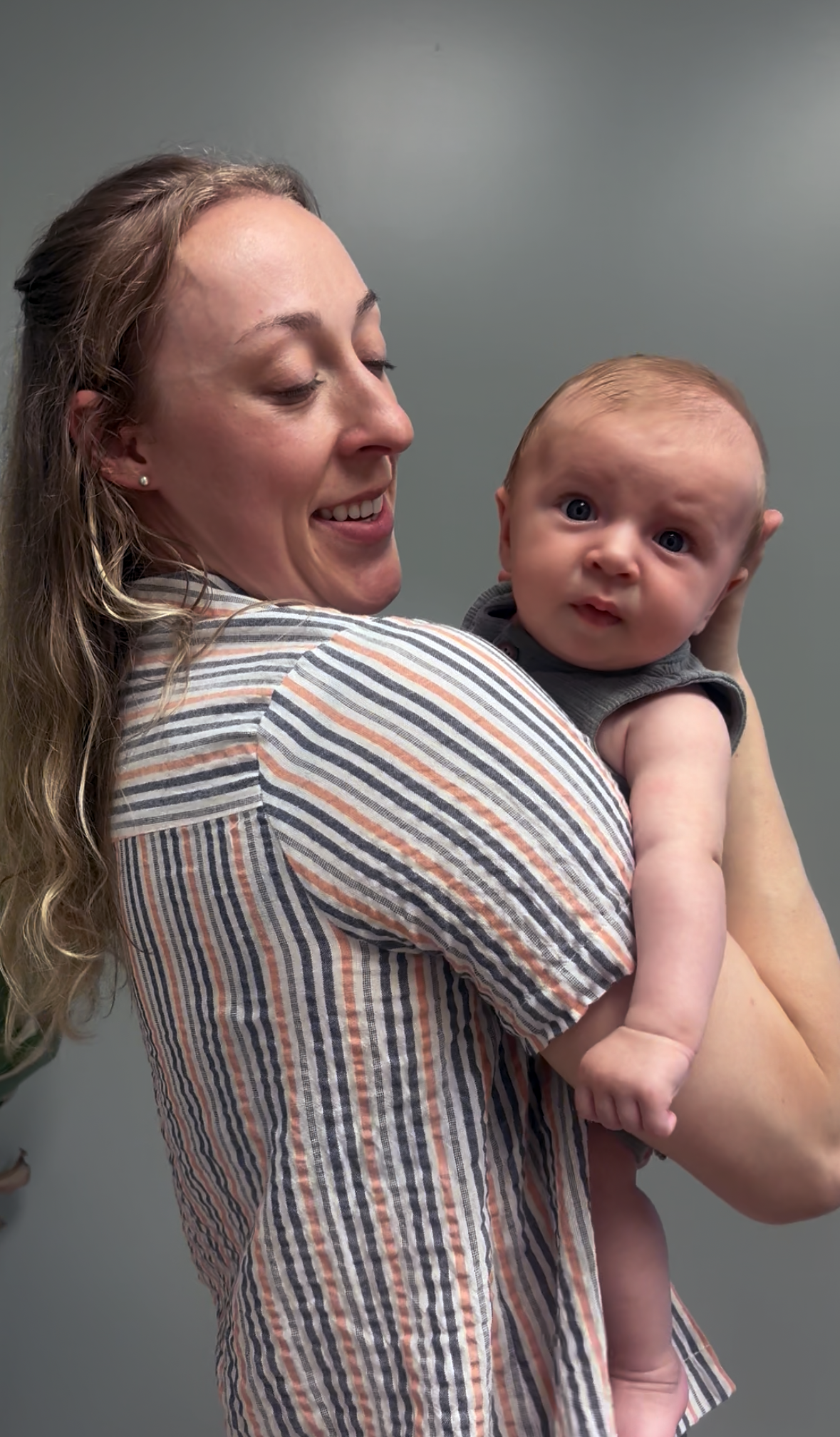 A woman with long wavy hair smiling and holding a baby with blue eyes. The woman is wearing a striped shirt and earrings, and the baby is in a sleeveless gray top. They are standing against a plain gray background.