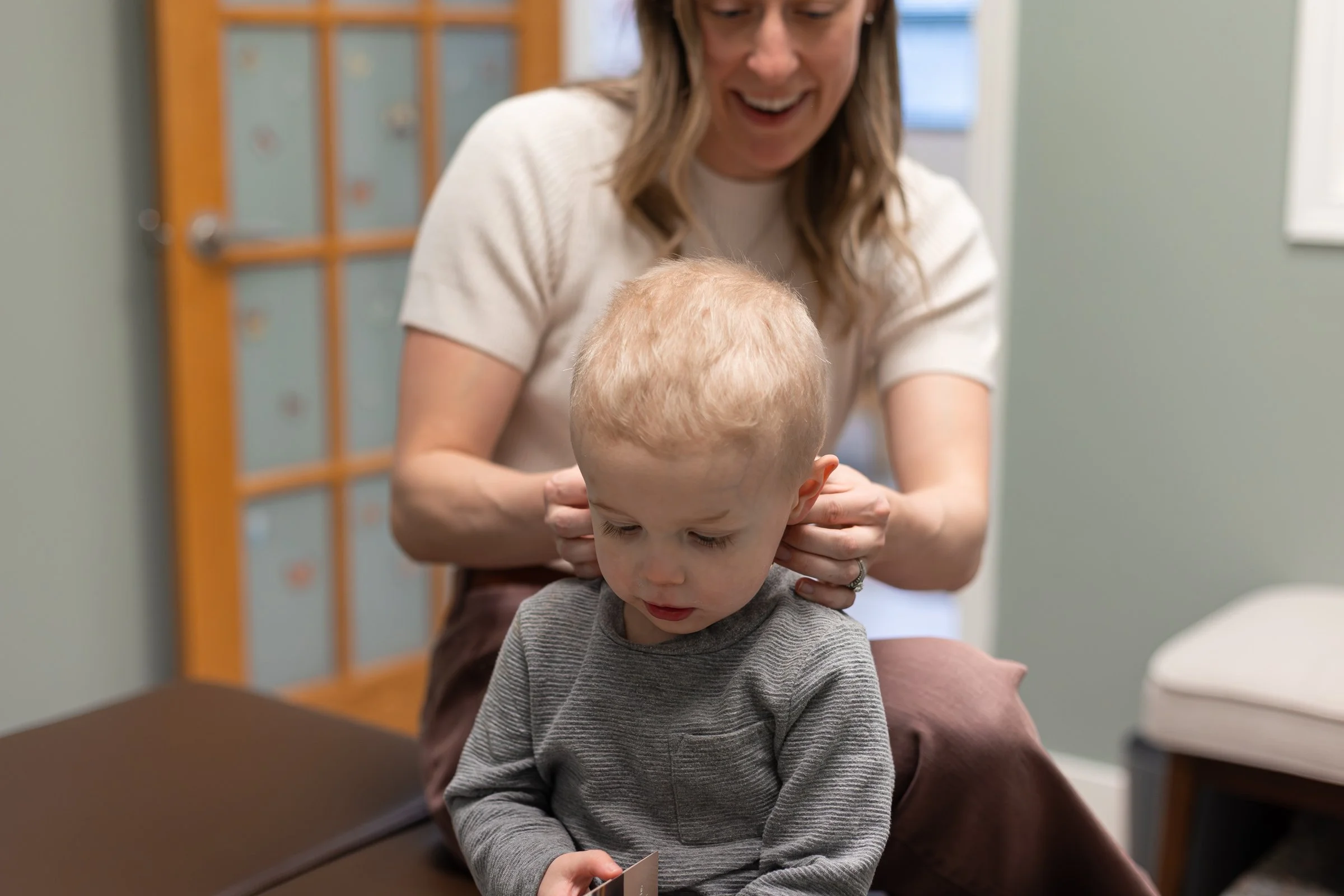 A woman with glasses smiling while gently holding a young boy's shoulders in a room with a wooden cabinet in the background.