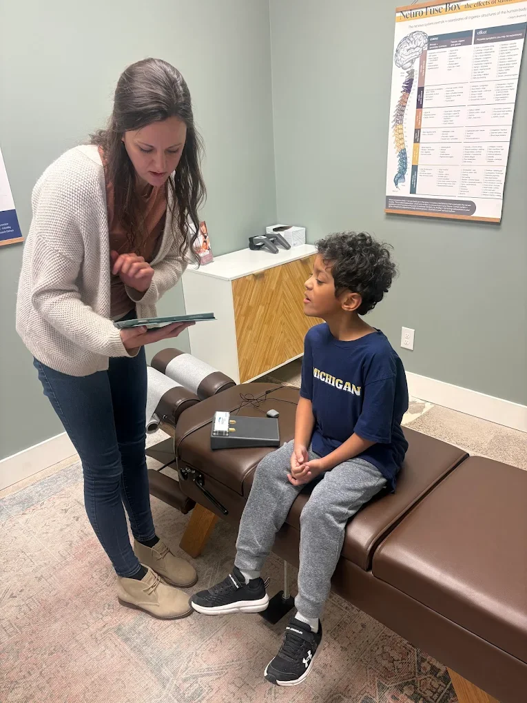 A young boy with curly hair and wearing a navy blue 'MICHIGAN' t-shirt sits on an examination table in a medical office. A woman, possibly a doctor or nurse, leans over him holding a clipboard and engaging in conversation. The room has a poster about the nervous system on the wall and a small cabinet with medical equipment.