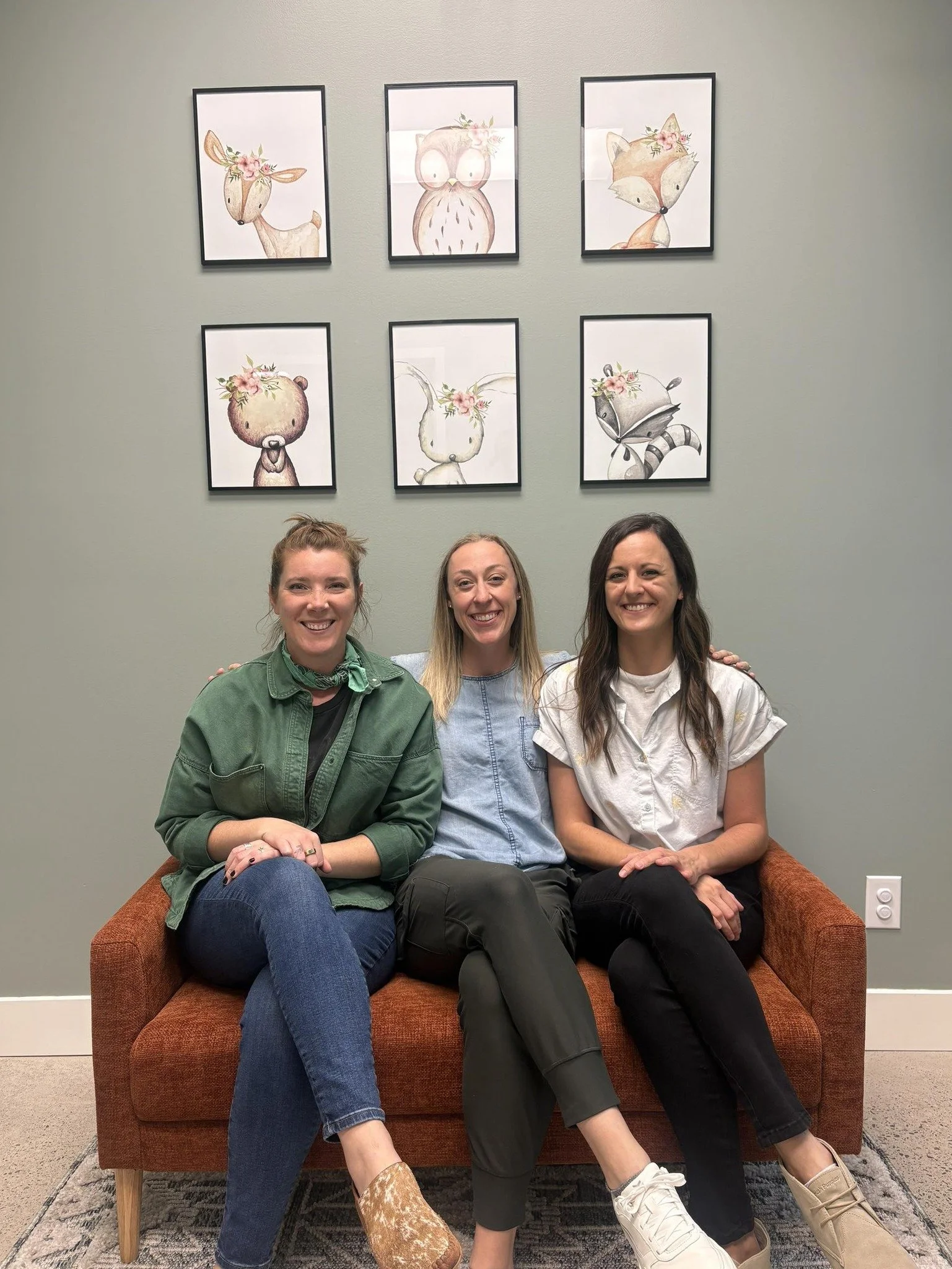 Three women sitting on an orange couch in front of a light green wall decorated with six framed, whimsical animal watercolor illustrations, smiling at the camera.