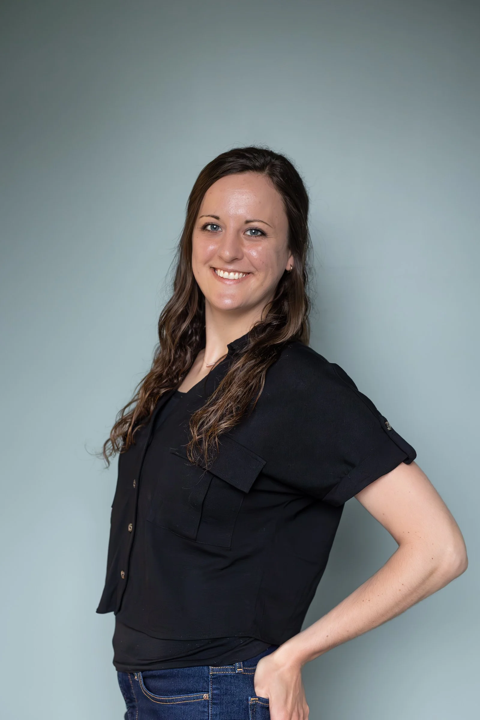A woman smiling and wearing a black shirt with rolled sleeves, standing against a plain light gray background.