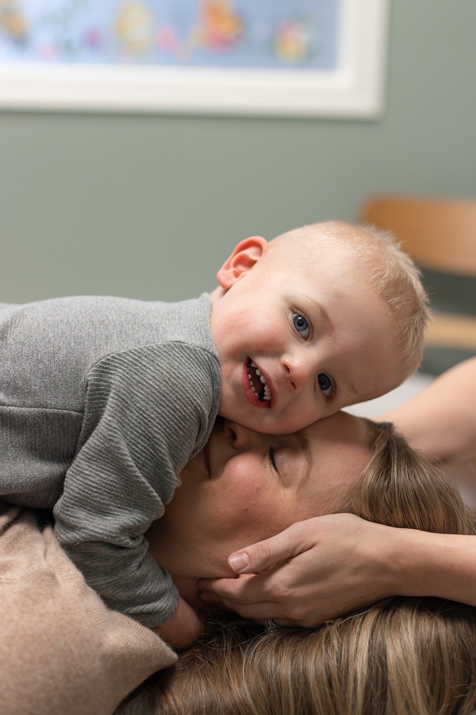 A young child with blonde hair and blue eyes smiling and lying on an adult's chest while being hugged in a tight embrace indoors.