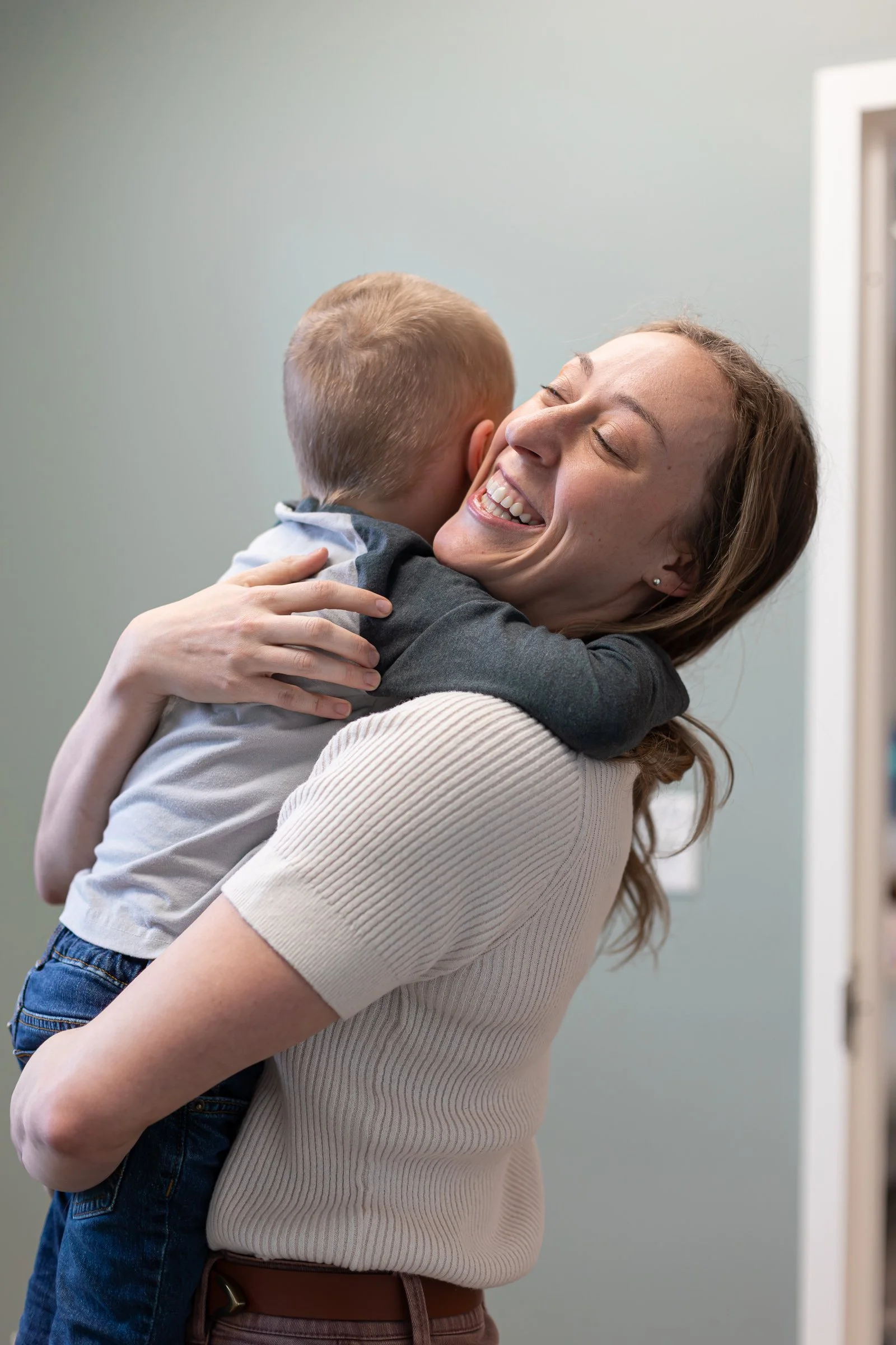 A woman with curly brown hair wearing a beige ribbed short sleeve top hugging a young boy, who has short blond hair and is wearing a gray T-shirt and blue jeans, both smiling with their eyes closed.