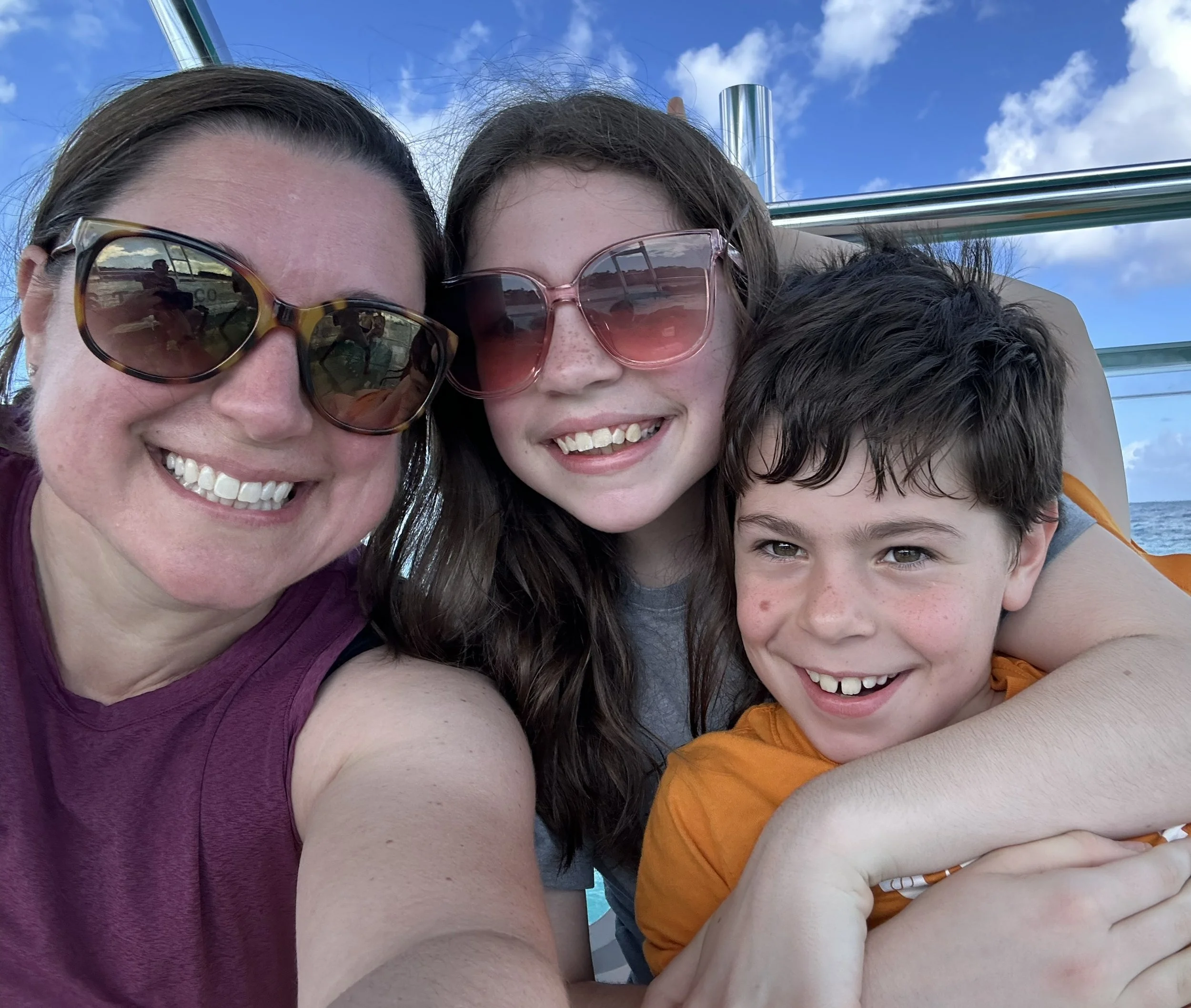 A smiling woman and two children wearing sunglasses on a boat with the ocean and blue sky in the background.