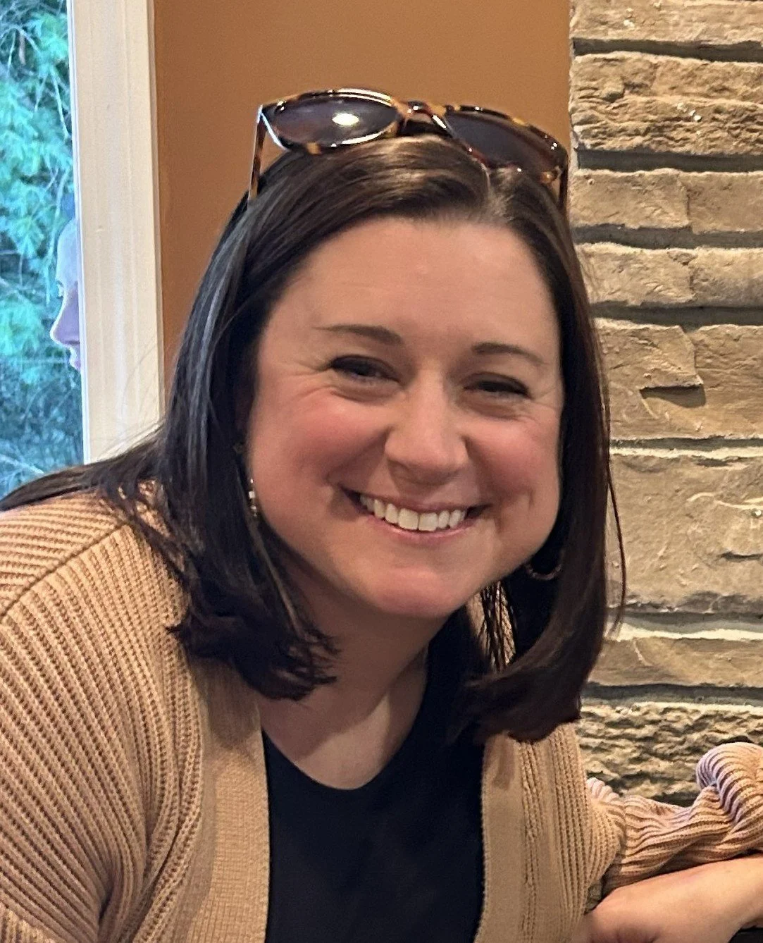 Smiling woman with dark brown hair, wearing sunglasses on her head, a black top, and a tan cardigan, sitting indoors near a stone wall and a window with greenery outside.