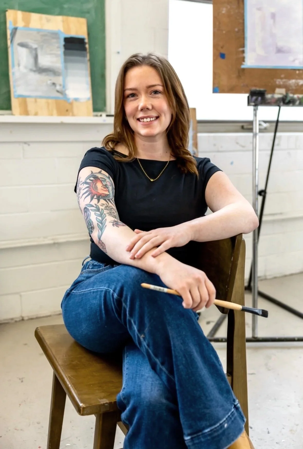 A woman with shoulder-length brown hair, tattooed arm, wearing a black t-shirt, blue jeans, and a gold necklace, sits on a wooden chair holding a paintbrush, smiling in an art studio with paintings and art supplies in the background.
