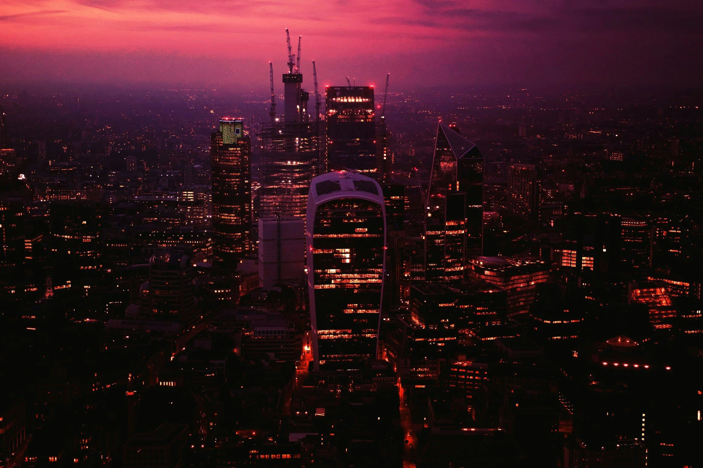 Nighttime cityscape of a downtown area with illuminated skyscrapers and the iconic London Eye Ferris wheel, with a pink and purple sunset sky in the background.