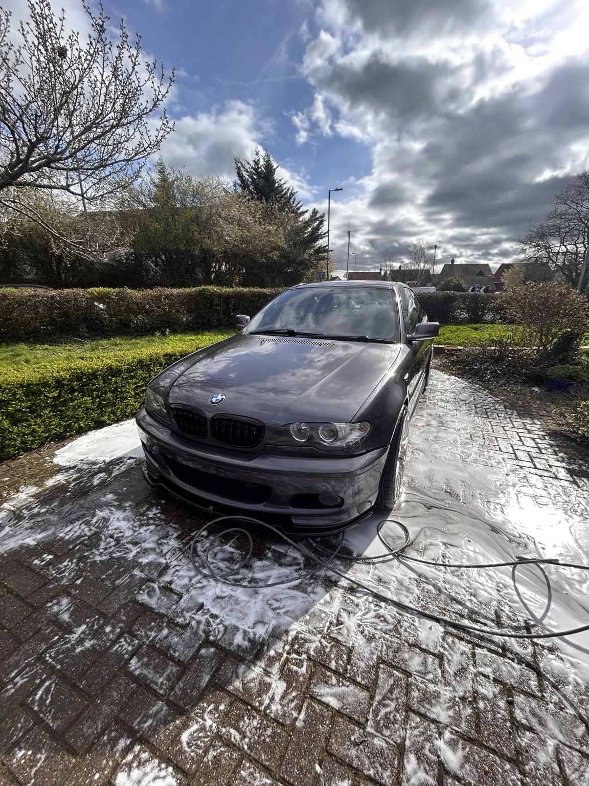 Black BMW car being washed outdoors on a brick driveway with soap suds, soap, and hoses visible. In the background, there are trees, bushes, and a partly cloudy sky.