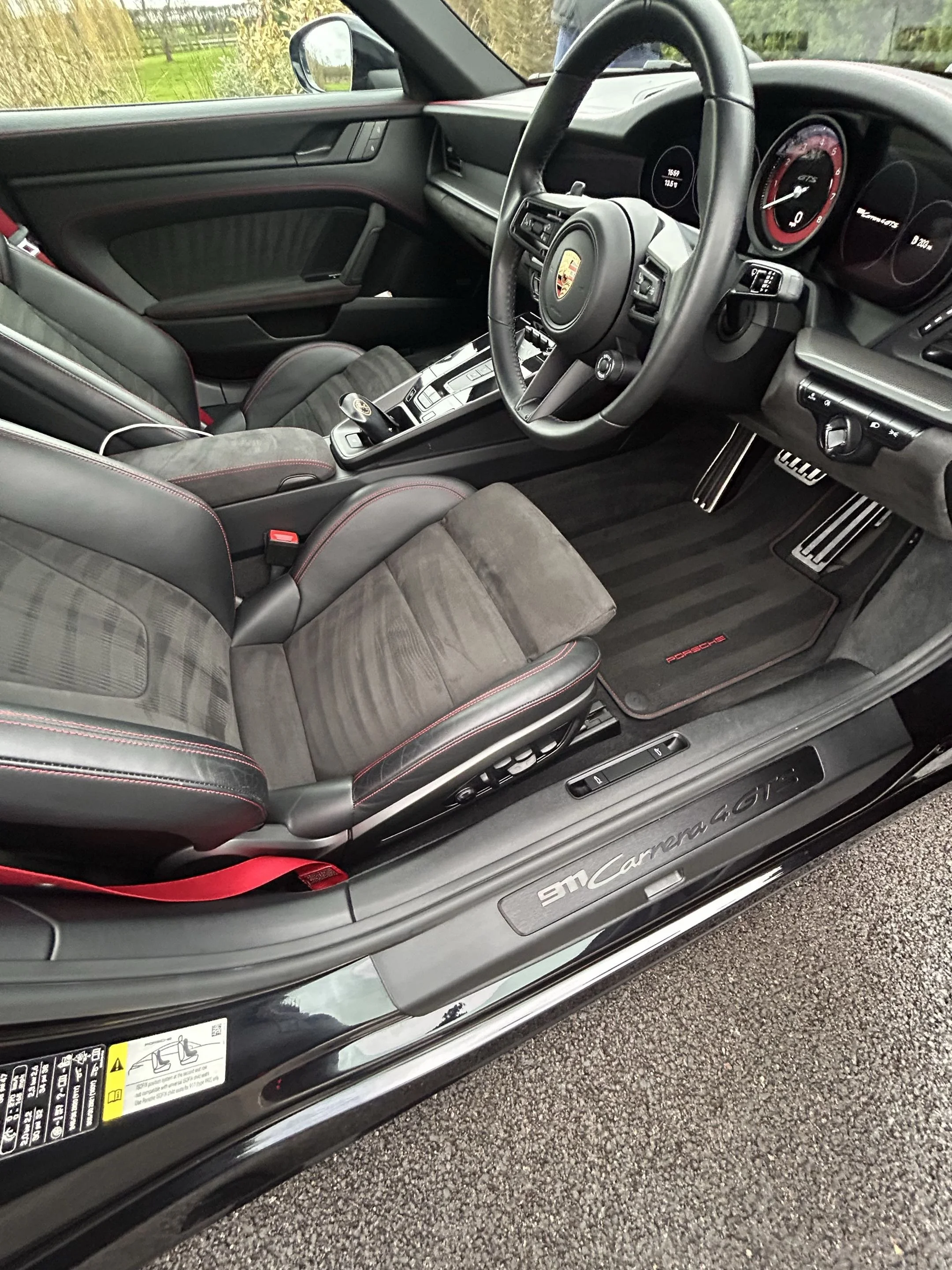 Interior of a black Porsche 911 Carrera GTS, showing black leather and Alcantara seats with red stitching, the steering wheel with the Porsche emblem, and the dashboard with analog gauges and digital displays.