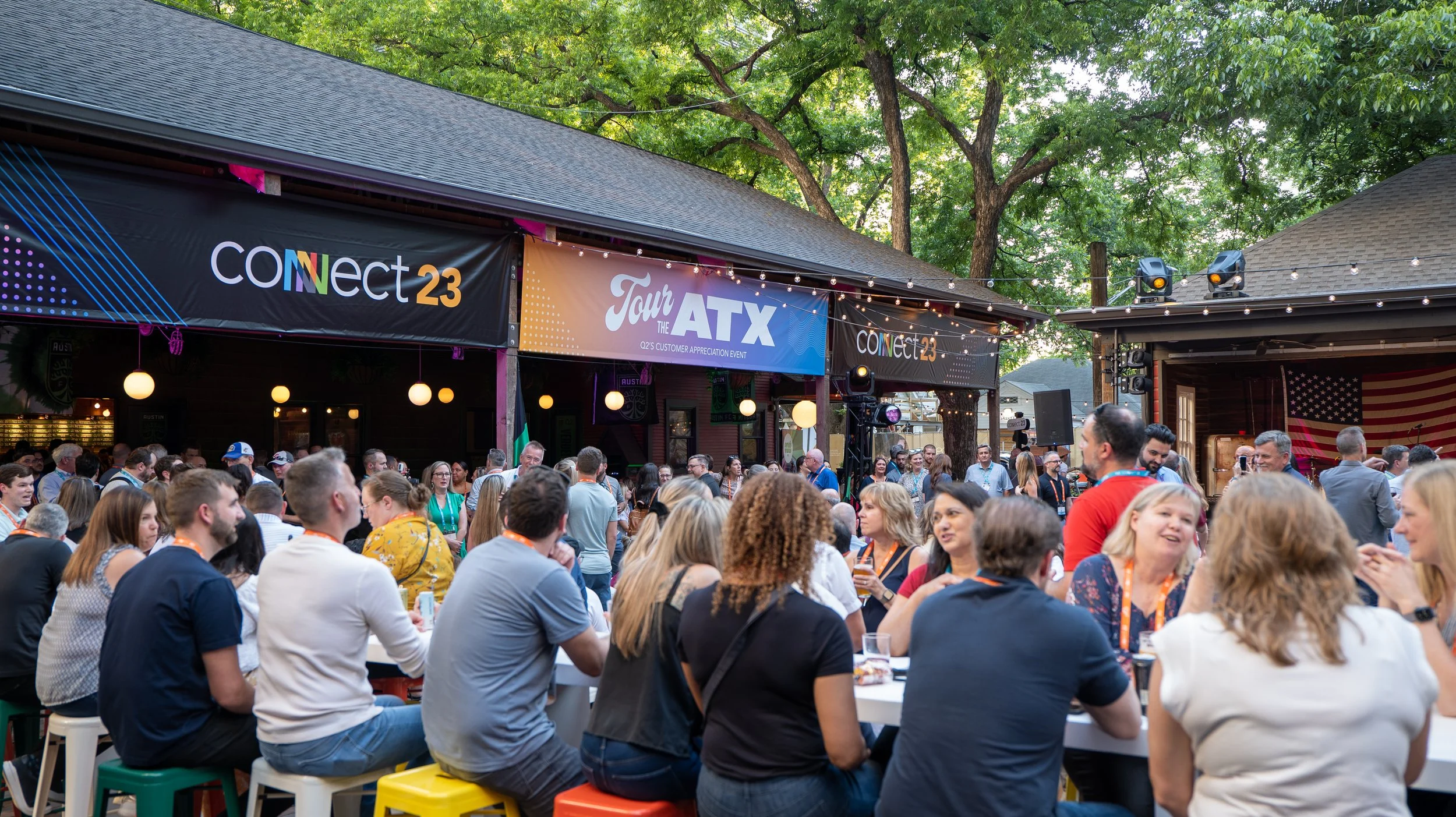 A large outdoor gathering of people at a social event with banners that read 'connect 23' and 'Tour the ATX.' People are seated and standing, engaging in conversations, with a stage and sound equipment visible. The setting is in a shaded area with trees and string lights overhead.