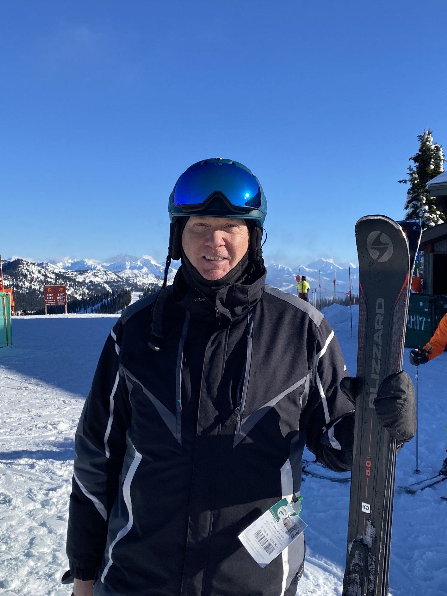 A man wearing a black ski jacket and a blue helmet with dark goggles, standing on snow with mountains in the background, holding a pair of skis.