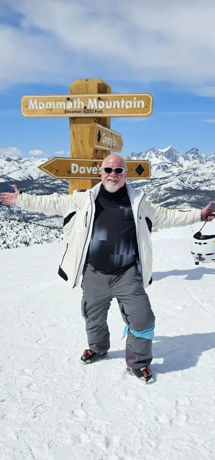 A man in winter gear standing on snow with arms outstretched, smiling, in front of a signpost at Mammoth Mountain, with snowy mountains in the background.