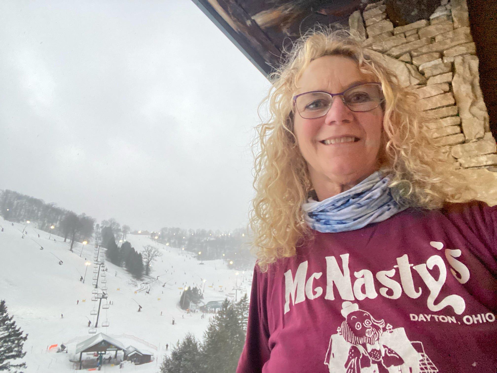 Kittyhawk Ski Club member.  A woman with curly blonde hair, glasses, and a purple 'MGNasty's Dayton, Ohio' T-shirt, smiling while taking a selfie at a snowy ski resort, with ski lifts and snow-covered slopes in the background.