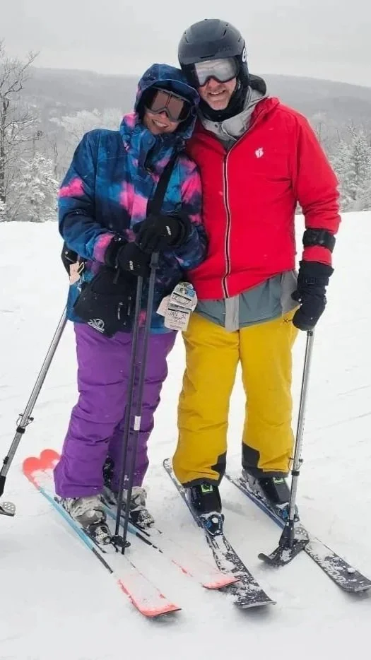 Two people wearing ski gear and helmets, standing on snow with skis, smiling together in a winter landscape.