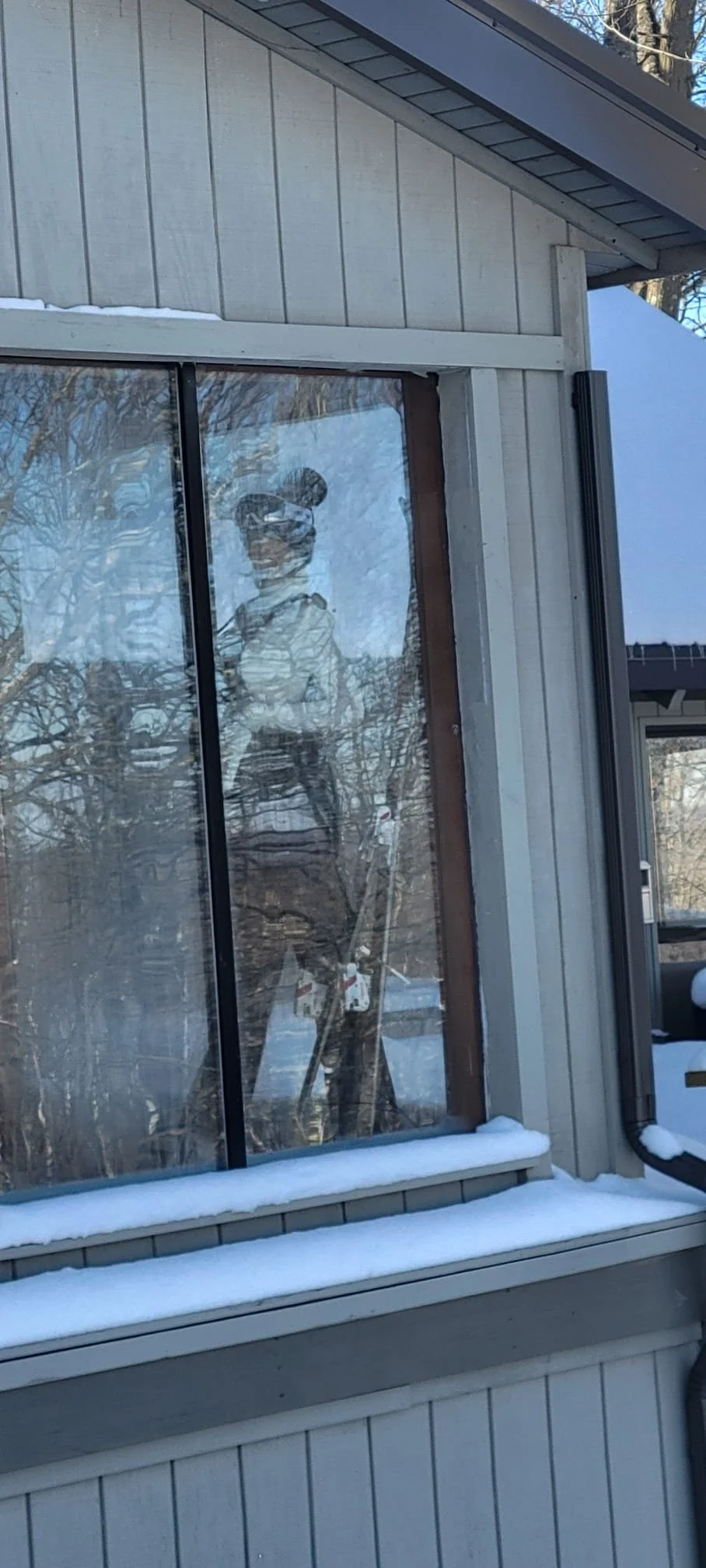 Kittyhawk Ski Club. Reflection of a woman in a glass window, wearing sunglasses and a baseball cap, standing outdoors with trees and snow in the background.