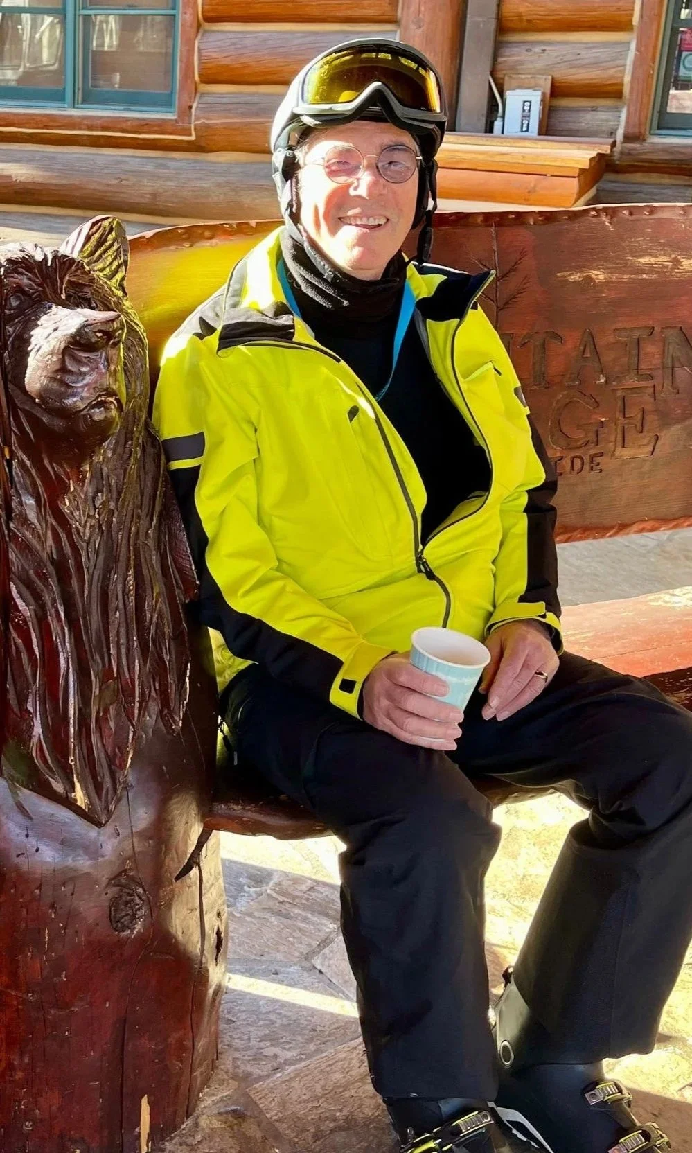 A man in ski gear, including a bright yellow jacket, black pants, ski boots, and ski helmet with goggles, sitting on a wooden bench with a carved bear figure and holding a coffee cup. The background shows a log cabin exterior.