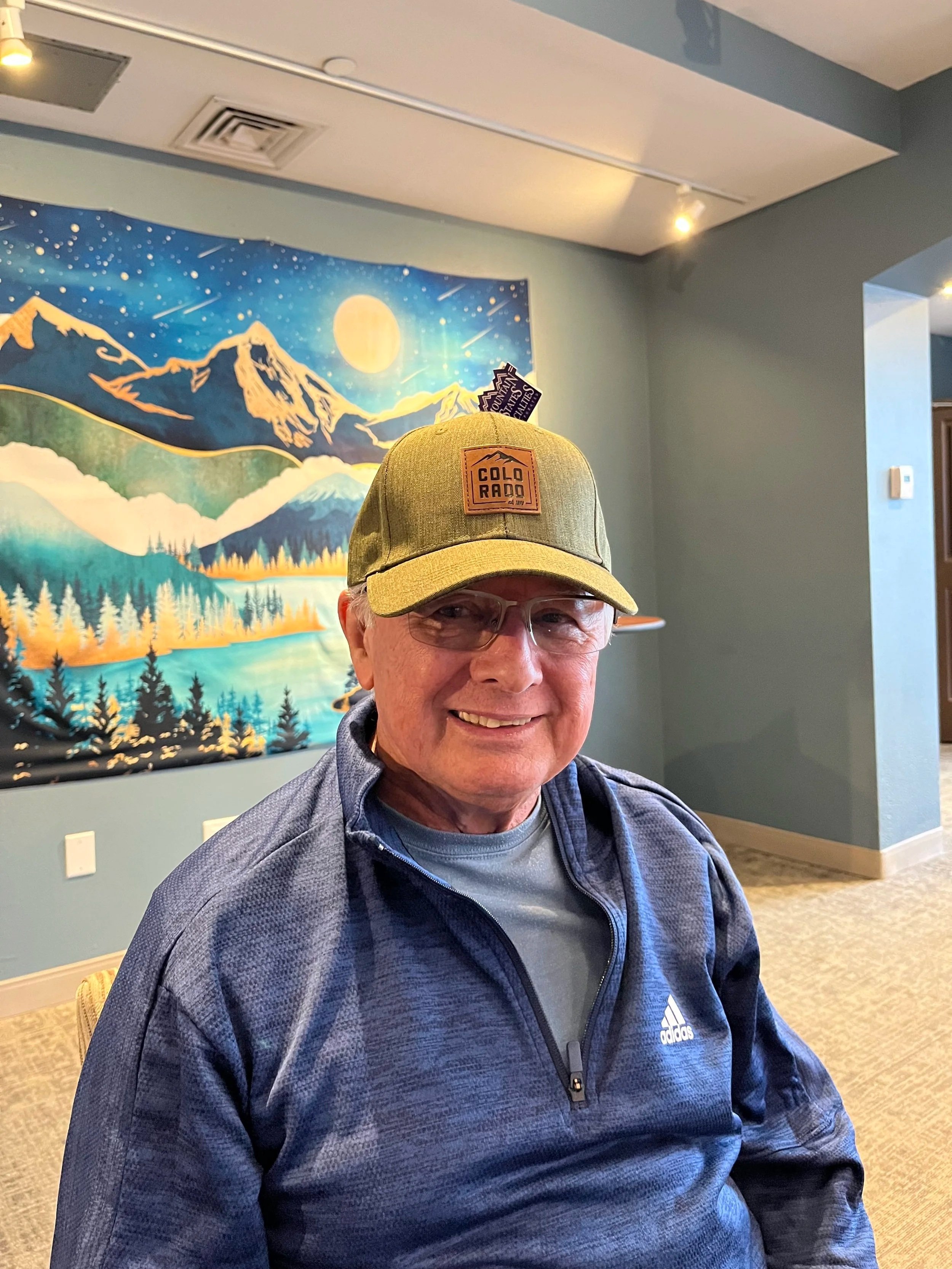 A smiling elderly man wearing glasses, a blue zip-up Adidas jacket, and a green baseball cap with a 'Cold Rado' patch, sitting indoors with a mountain landscape tapestry behind him.