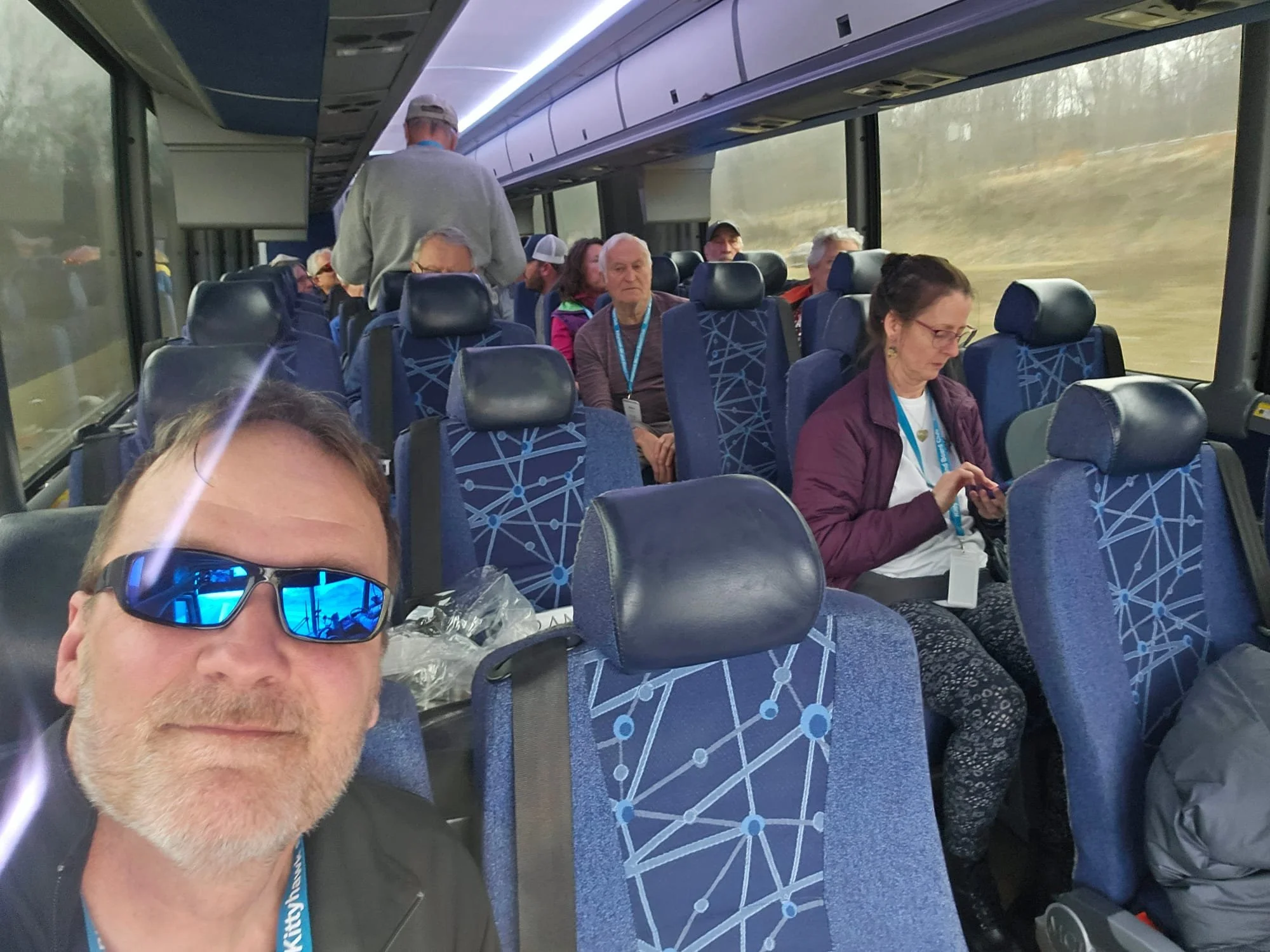 Kittyhawk Ski Club members.  A man with sunglasses taking a selfie onboard a bus with other passengers seated behind him, some looking at their phones, during daytime travel.