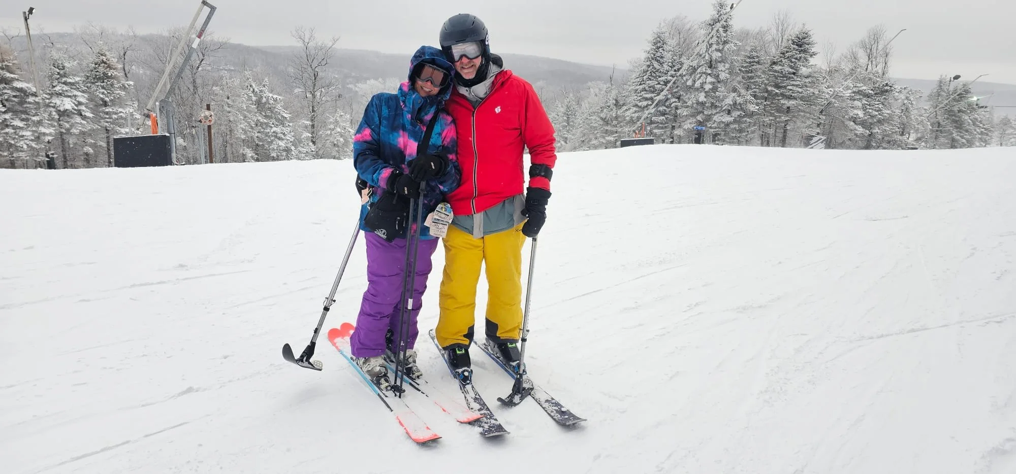 Kittyhawk Ski Club members.  Two people in colorful ski gear standing on snow with ski poles and skis, smiling, in a snowy landscape with trees and ski lift equipment in the background.