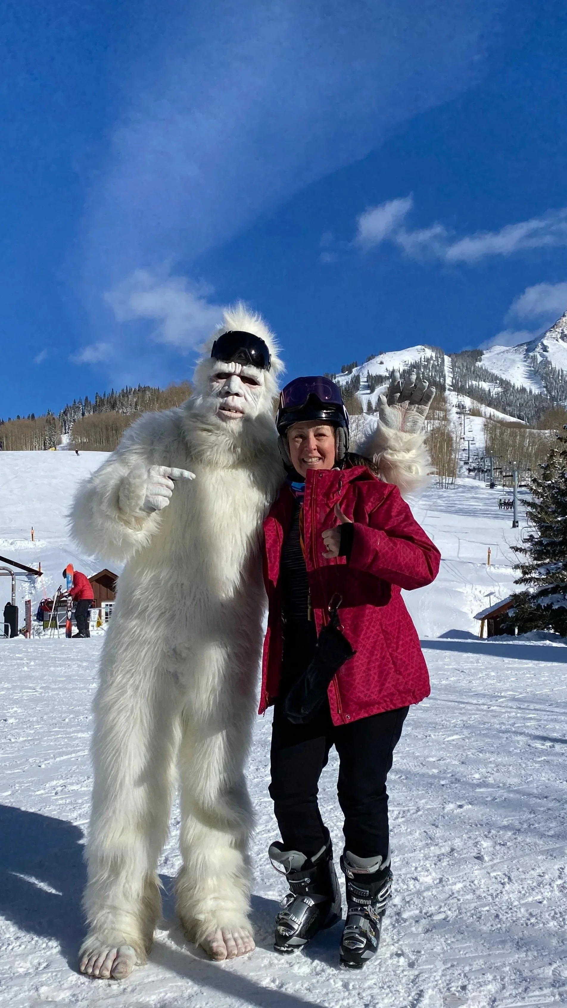 Person in pink ski jacket and black ski gear standing next to a person in a Sasquatch costume on a snowy mountain slope with ski lifts and trees in the background.