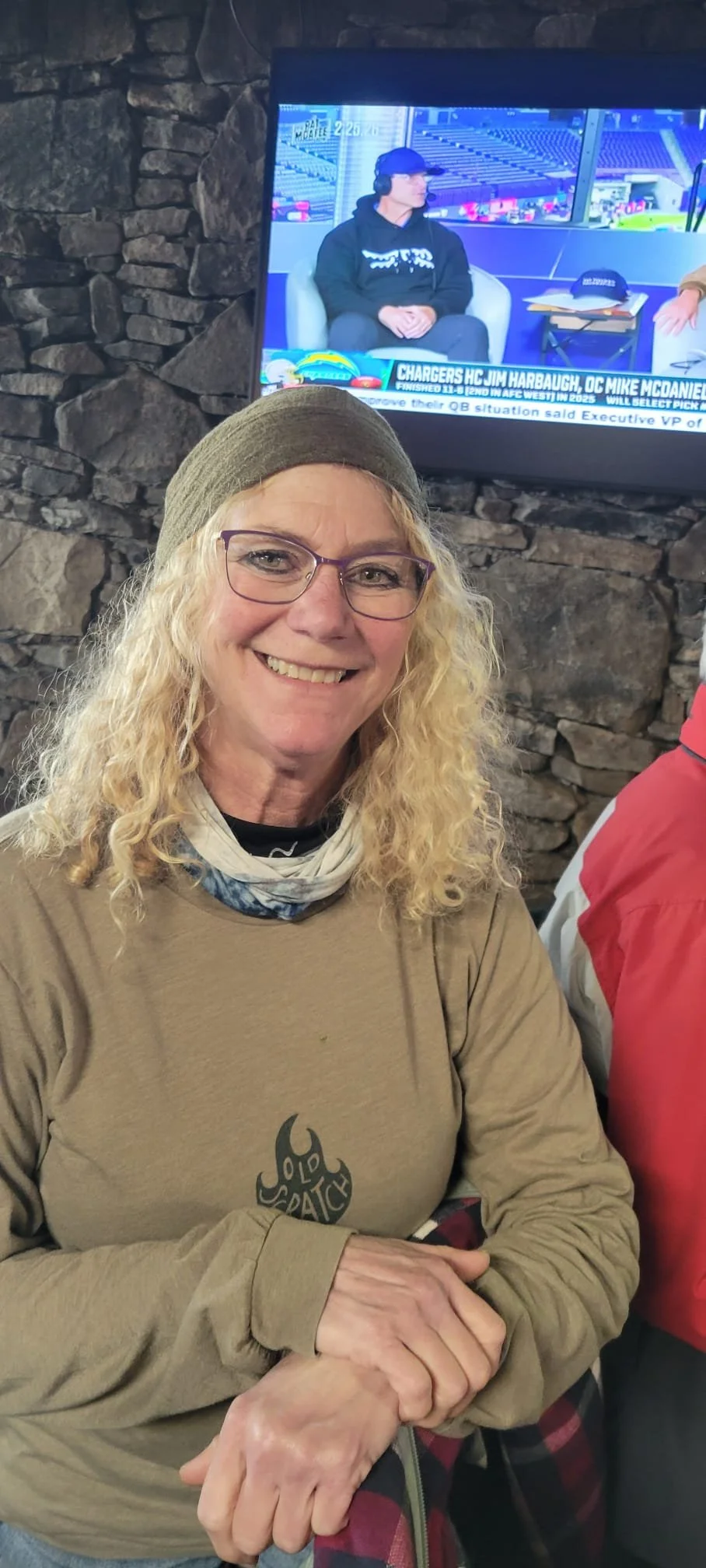 Kittyhawk Ski Club member.  A woman with long curly blonde hair, glasses, and a gray beanie, smiling, seated indoors in front of a stone wall with a television screen showing a sports broadcast.