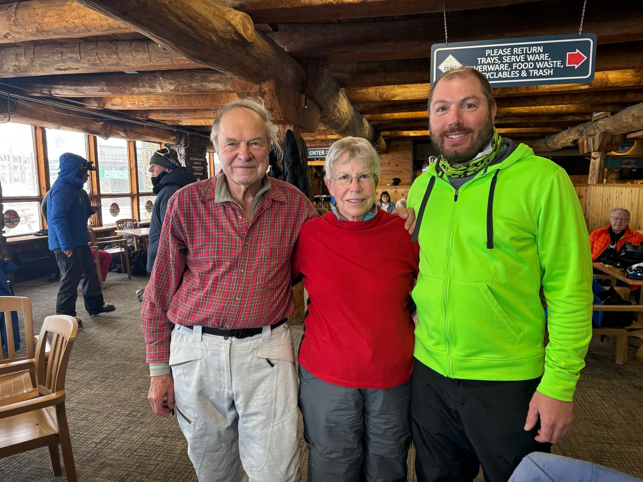 Kittyhawk Ski Club members.  Three people standing inside a rustic wooden lodge, smiling at the camera. An older man in a red plaid shirt, an older woman in a red sweater, and a young man in a bright green jacket.
