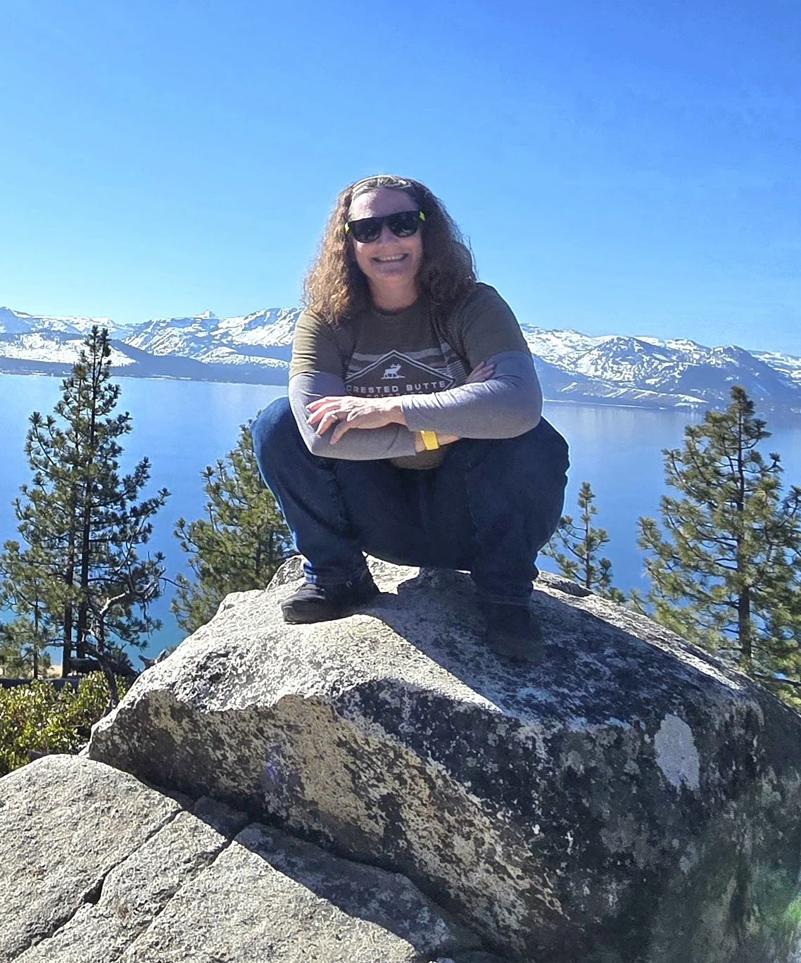 A woman squatting on a large rock in front of a lake with snow-capped mountains in the background, wearing sunglasses, a long-sleeved shirt, and black pants, smiling.