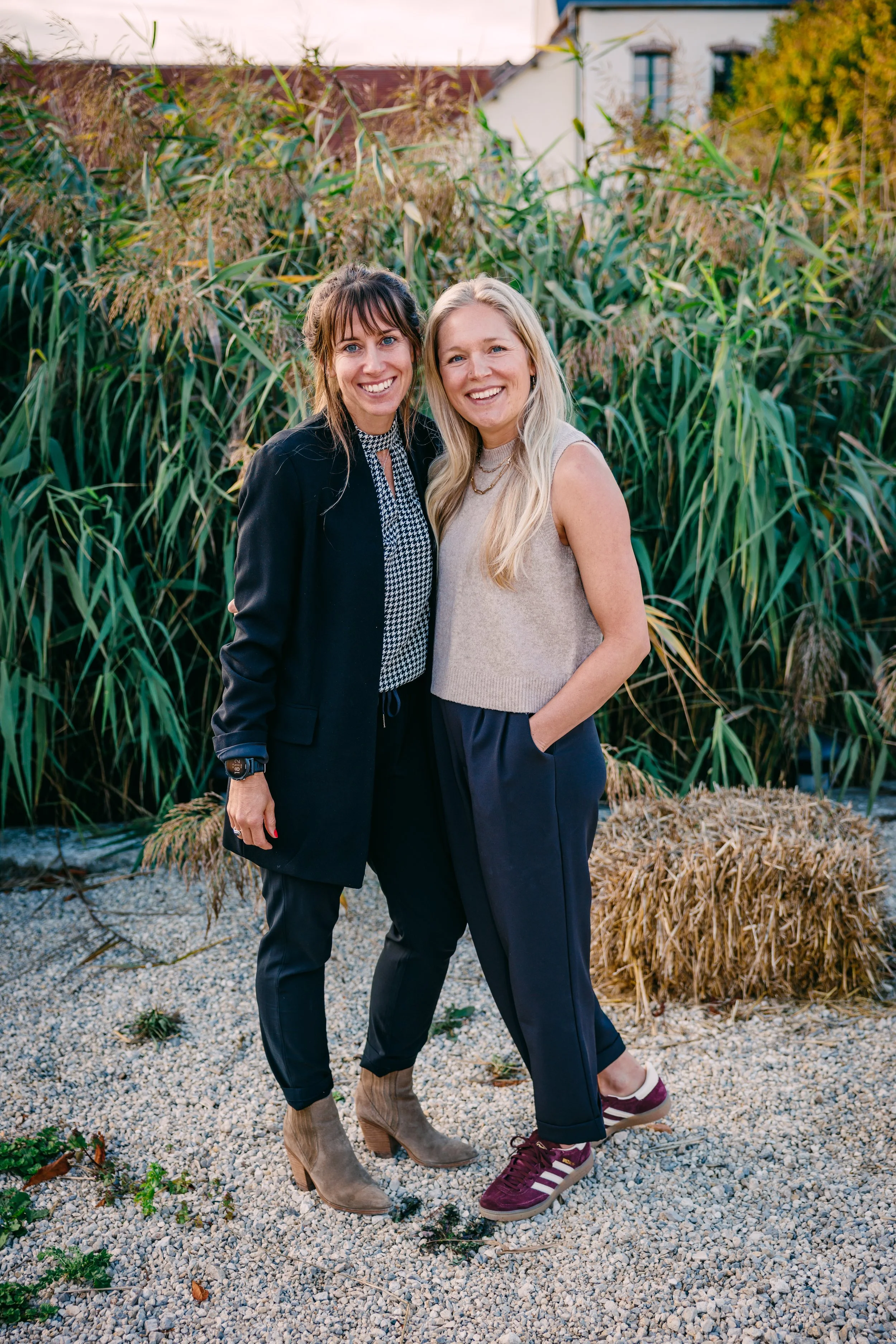 Two women standing outdoors in front of tall green plants, smiling at the camera. One woman has dark hair and is wearing a black blazer and patterned shirt, while the other has blonde hair and is dressed in a sleeveless beige top and dark pants. The setting appears to be a garden or natural area with a house visible in the background.
