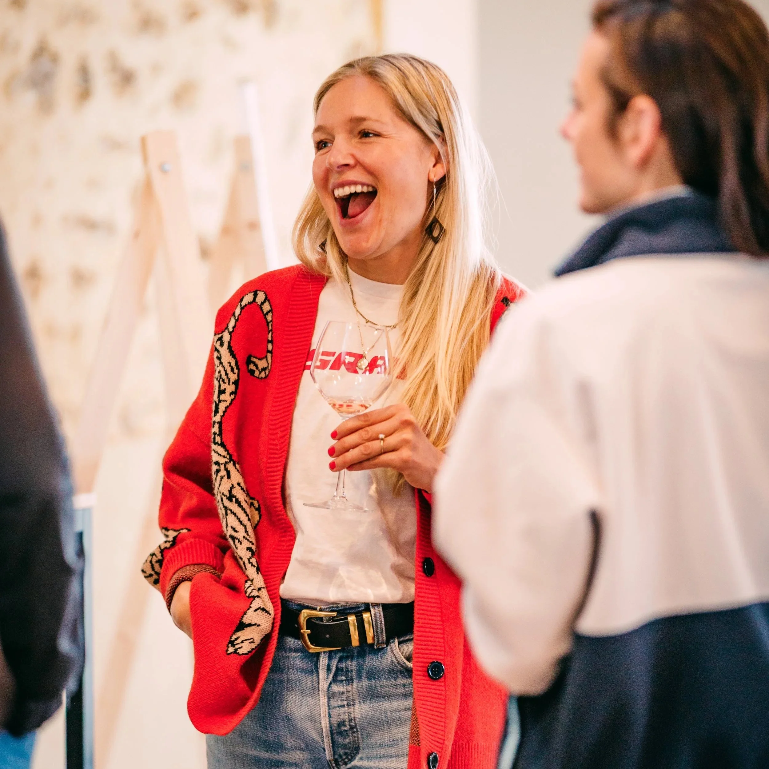 A woman with long blonde hair, smiling wearing a red cardigan with a leopard print design and a white t-shirt, standing in a social setting with other people.