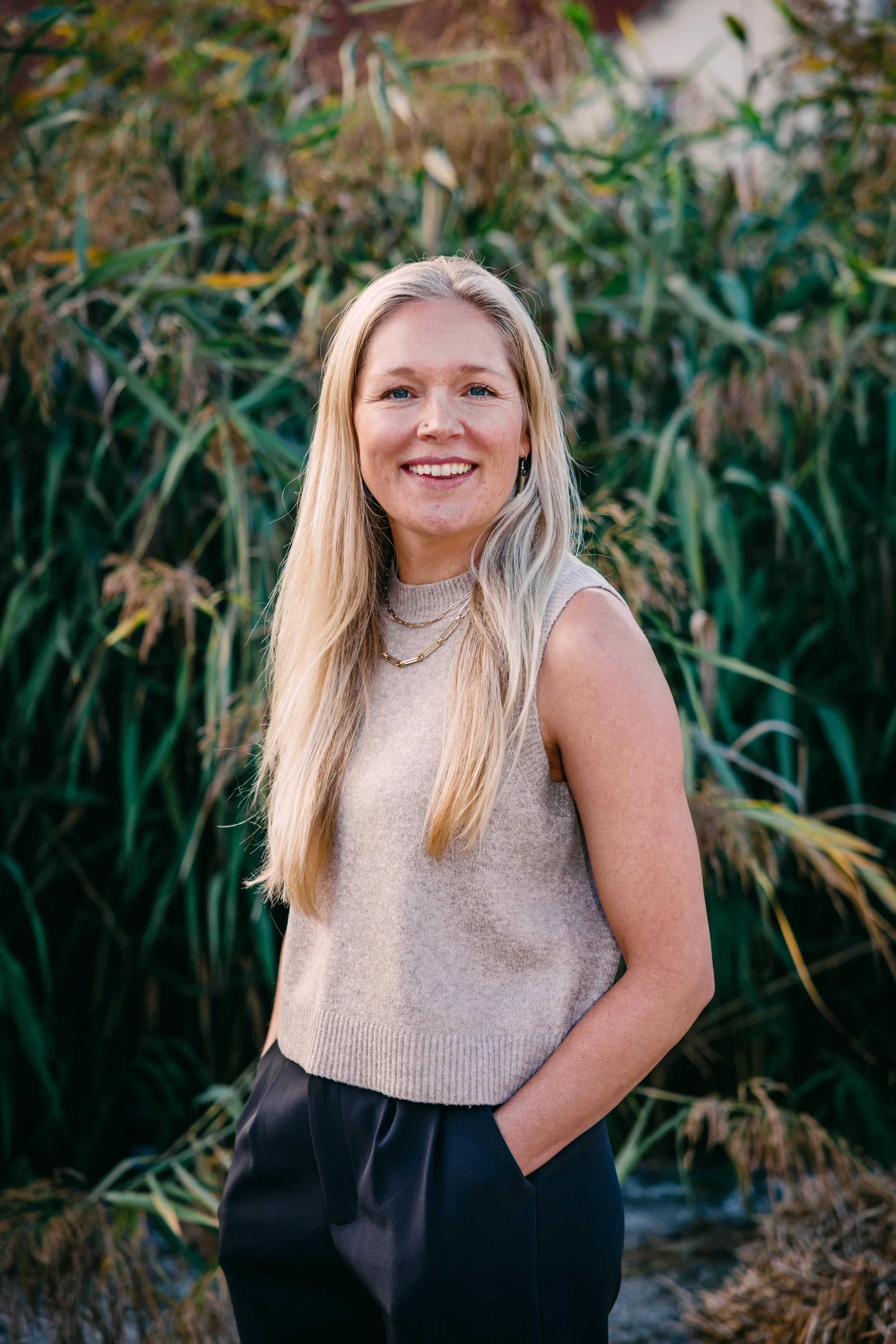 A woman with long blonde hair smiling outdoors in front of tall green grass and plants.