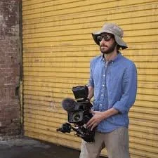 A man wearing a wide-brimmed hat, sunglasses, and a denim shirt holding a camera rig in front of a yellow metal door. This is Jeremy Emerman (Director of Photography).