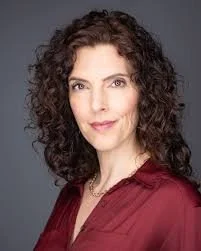 A woman with curly dark hair wearing a burgundy top looks at the camera against a gray background. This is Amy Levy.