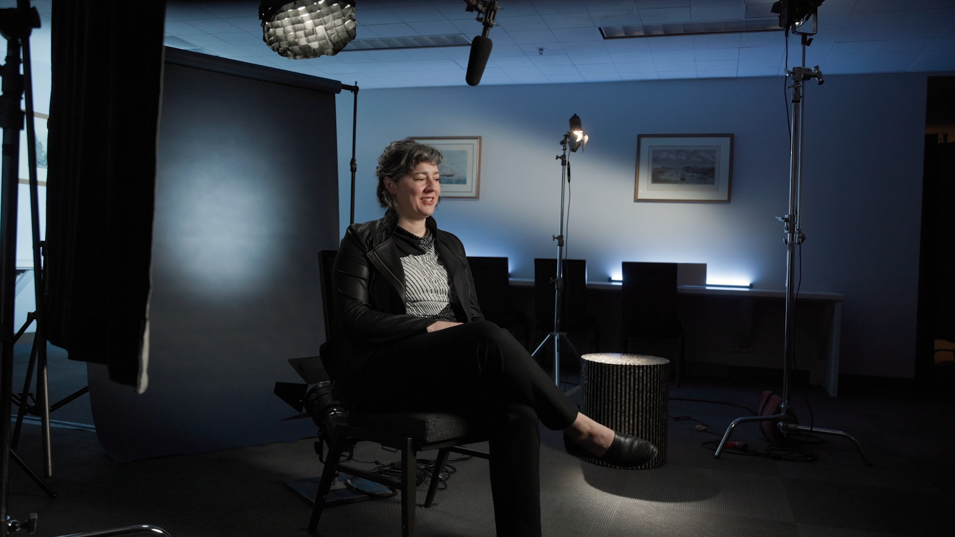 A woman sitting in a film studio, surrounded by professional lighting equipment, with a dark backdrop and framed pictures on the wall, during a video shoot of the Uncharted Territory documentary.