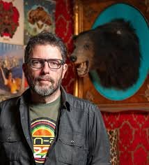 A man with glasses and a beard standing in front of colorful artwork and a mounted bear head. This is Charles de Lauzirika (Director).