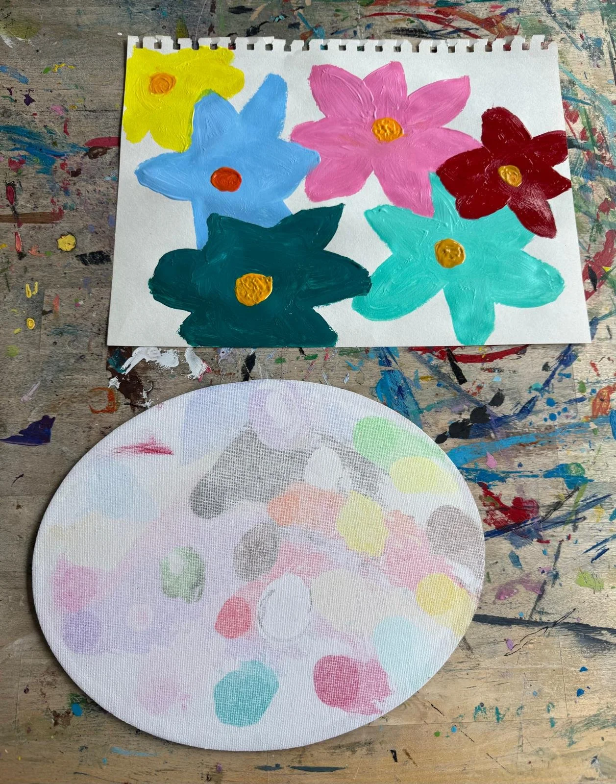 Square piece of paper with colorful painted flowers in pink, blue, red, yellow, and teal. A round piece of paper with pastel-colored polka dots set below the painted flowers, all on a splattered art table.